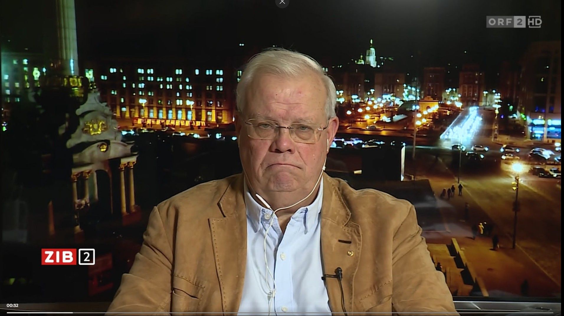 Ein älterer Mann mit Brille sitzt vor einer nächtlichen Stadtansicht im Fernsehstudio.