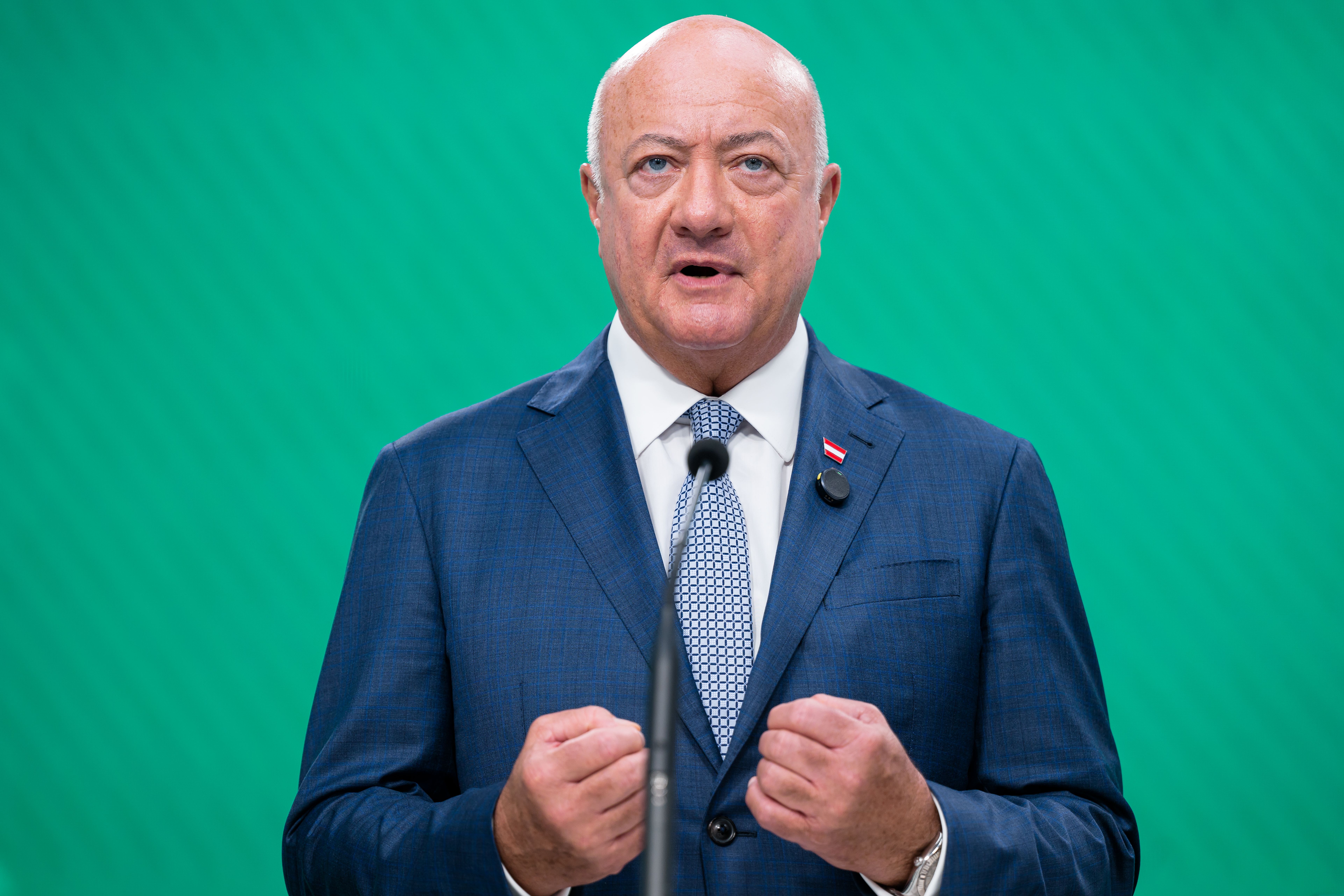 Ein Mann im blauen Anzug spricht bei einer Pressekonferenz vor grünem Hintergrund.