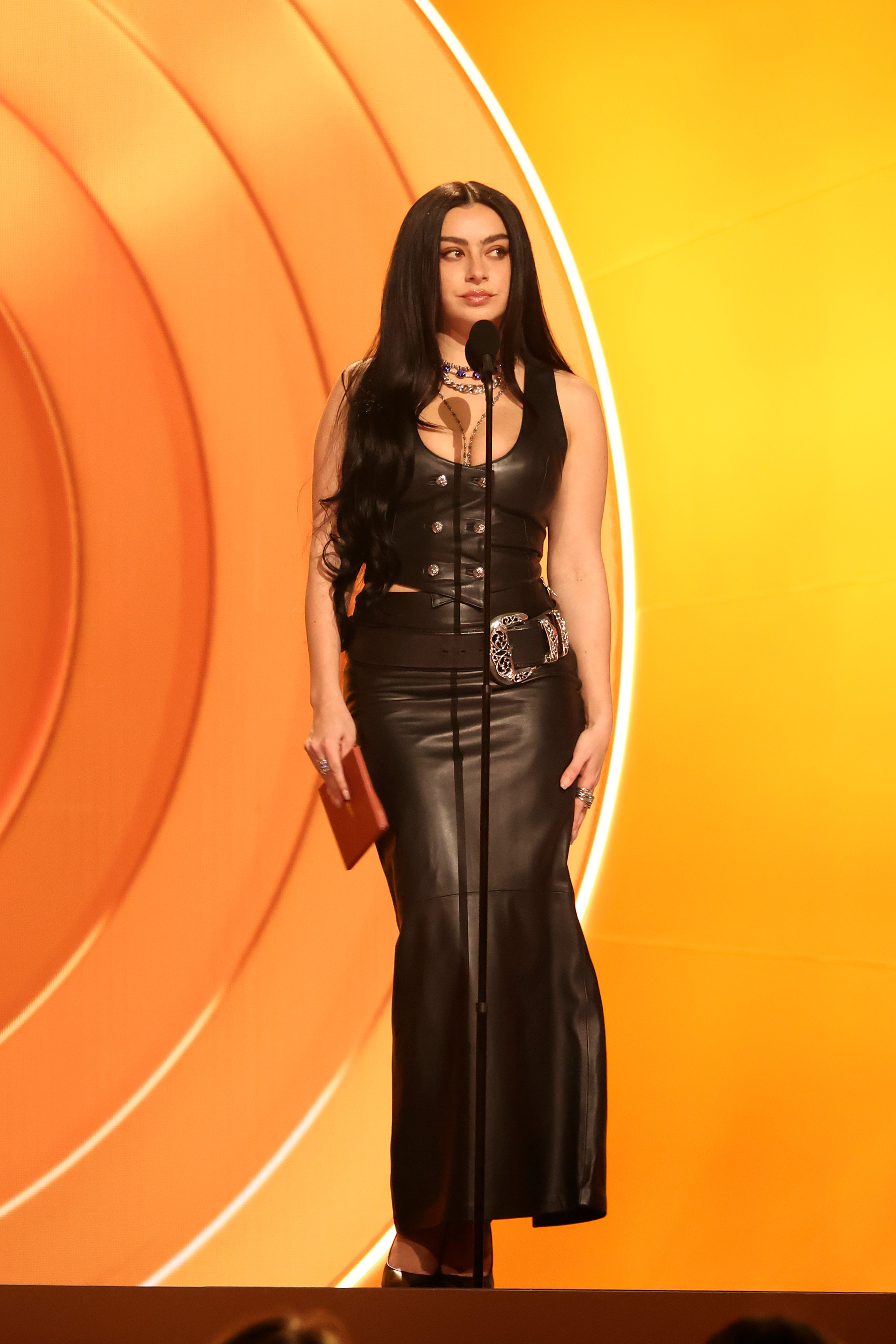 Frau im schwarzen Lederkleid spricht bei einer Preisverleihung auf orange-gelbem Hintergrund.