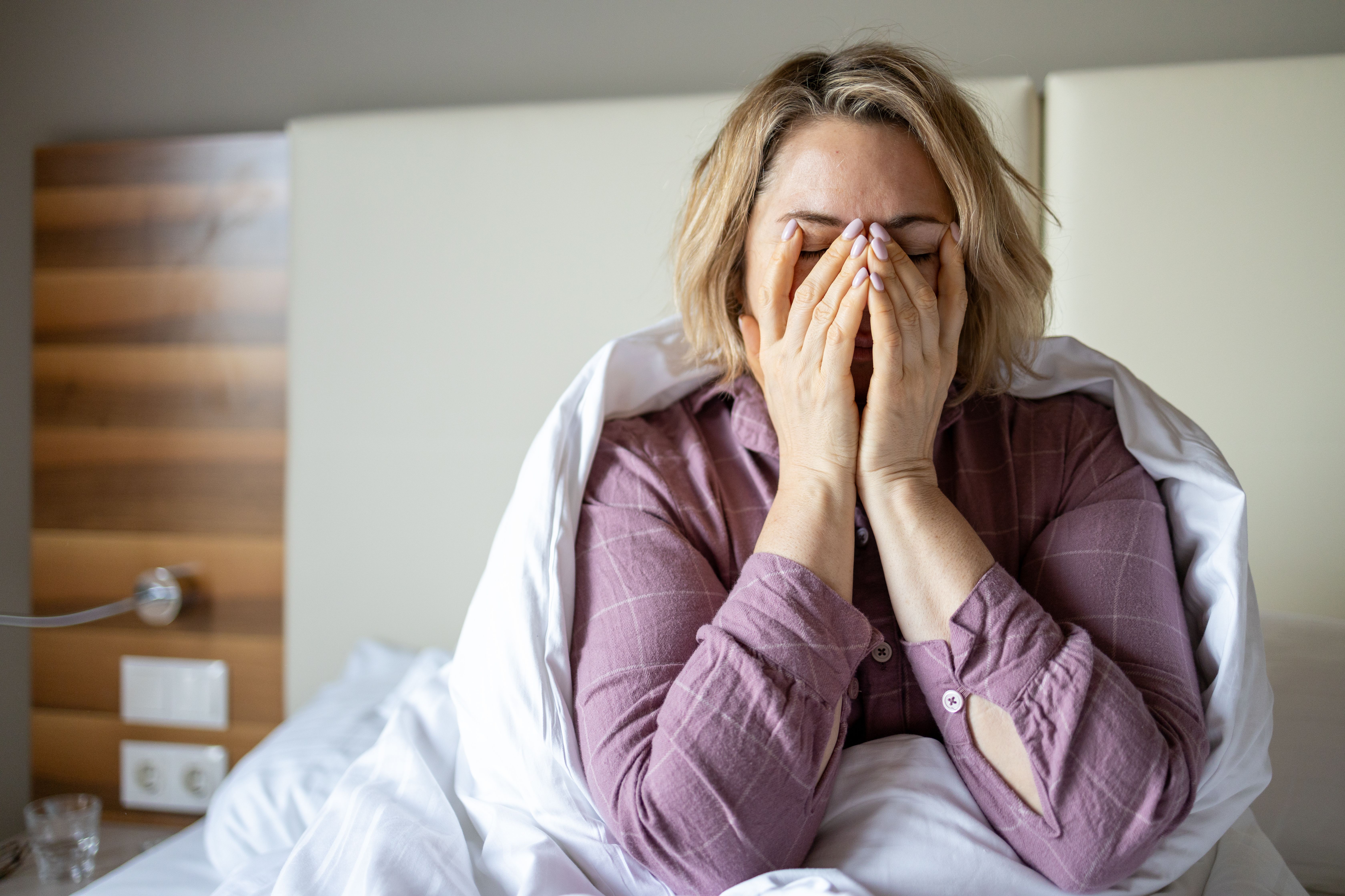 Frau sitzt im Bett, hält das Gesicht in den Händen und wirkt verzweifelt oder traurig.