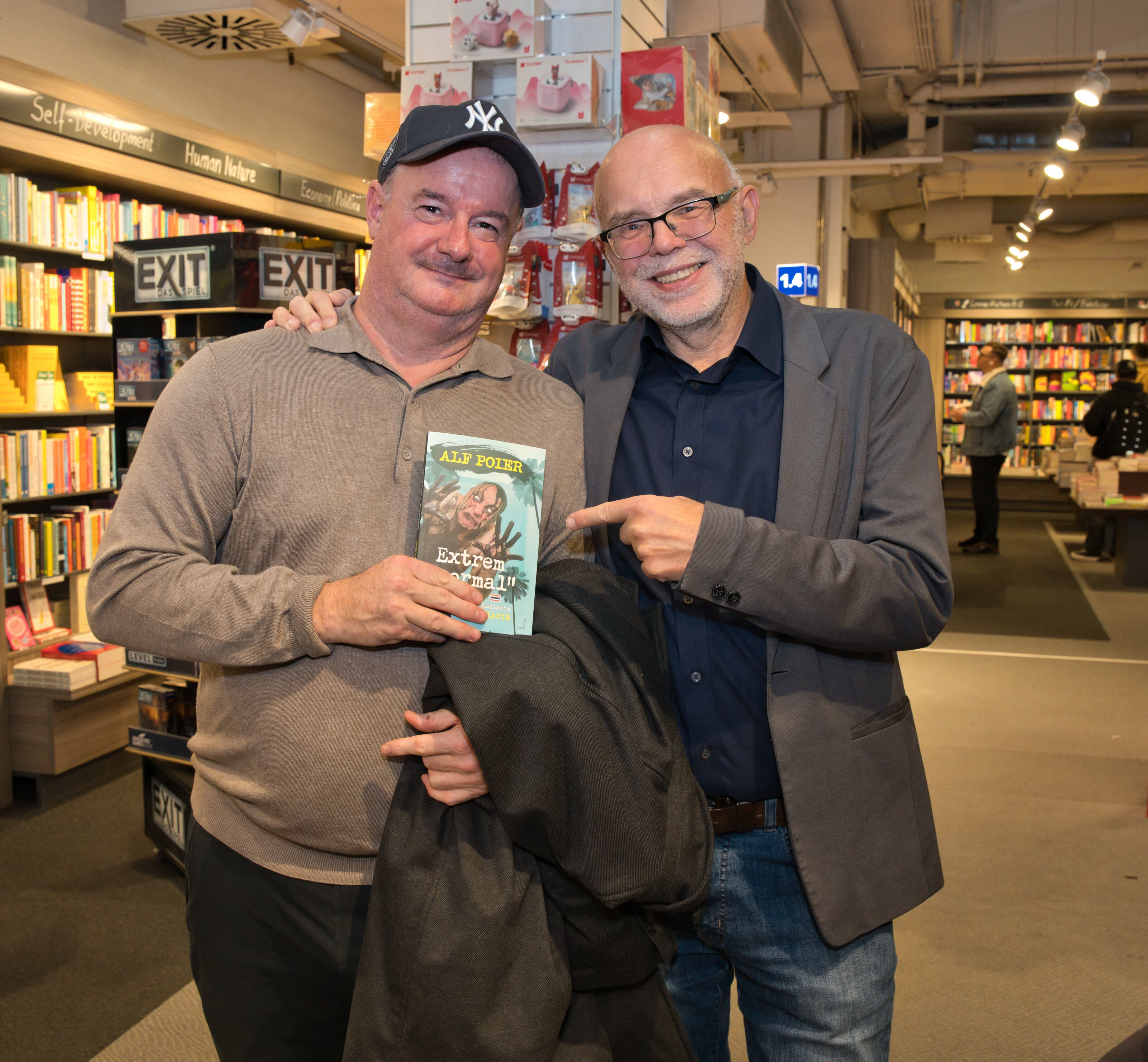 Zwei Männer in einer Buchhandlung, einer hält das Buch „Extrem normal!“ von Alf Poier.