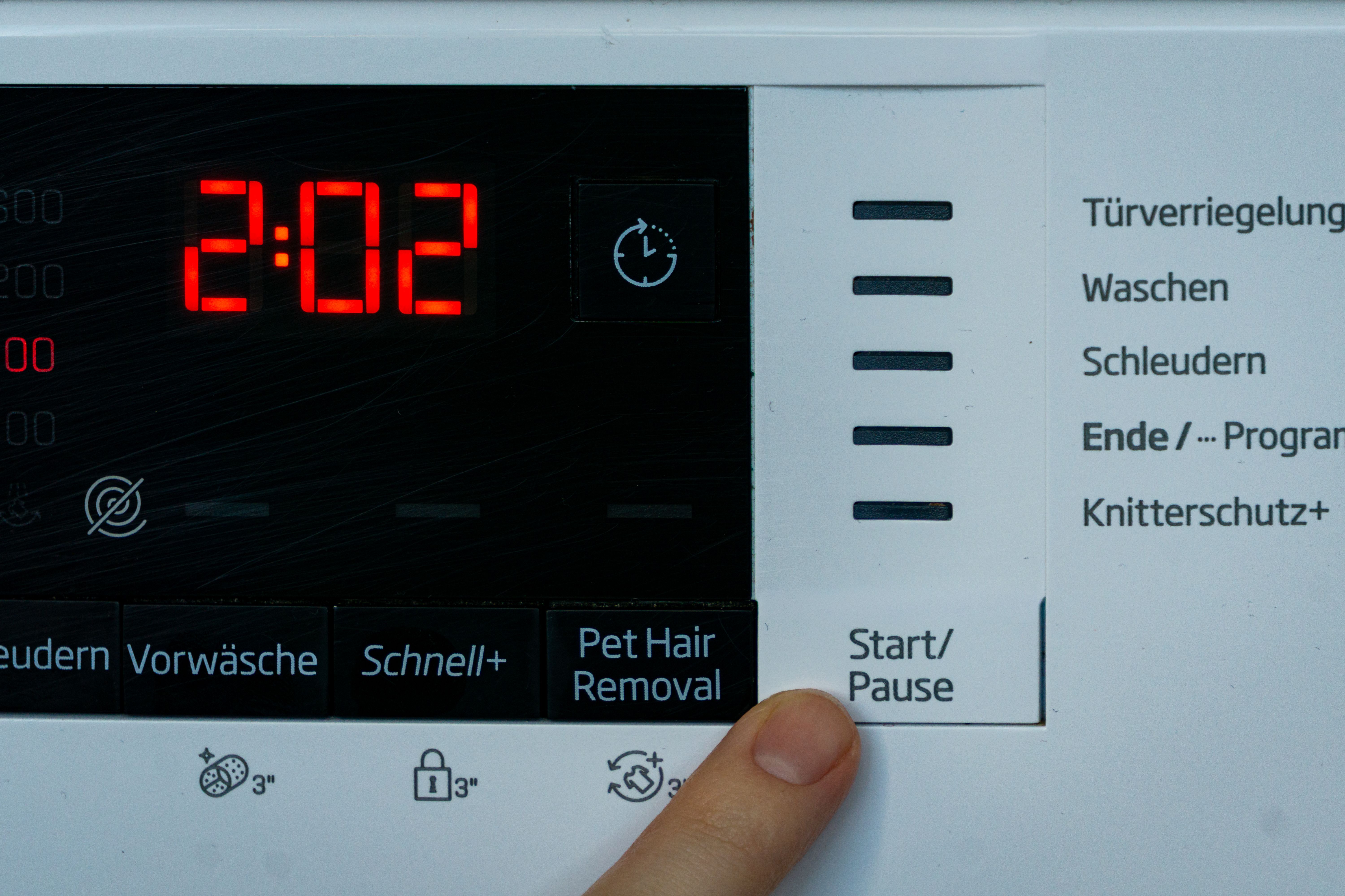 Finger drückt die Start/Pause-Taste an einer Waschmaschine mit digitalem Timer auf 2:02 Minuten.