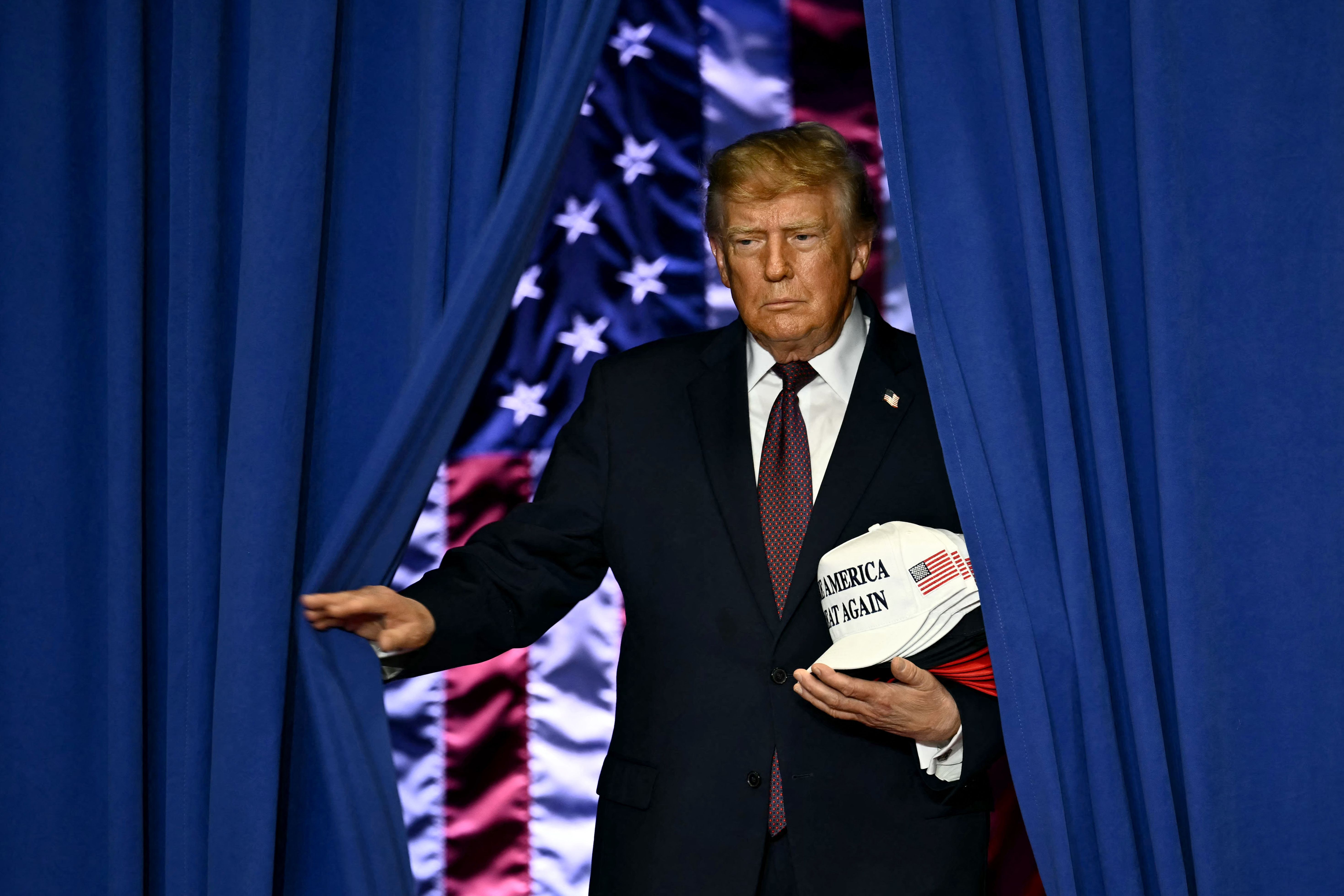 Ein Mann im Anzug tritt durch blaue Vorhänge vor eine US-Flagge und hält eine weiße Kappe.