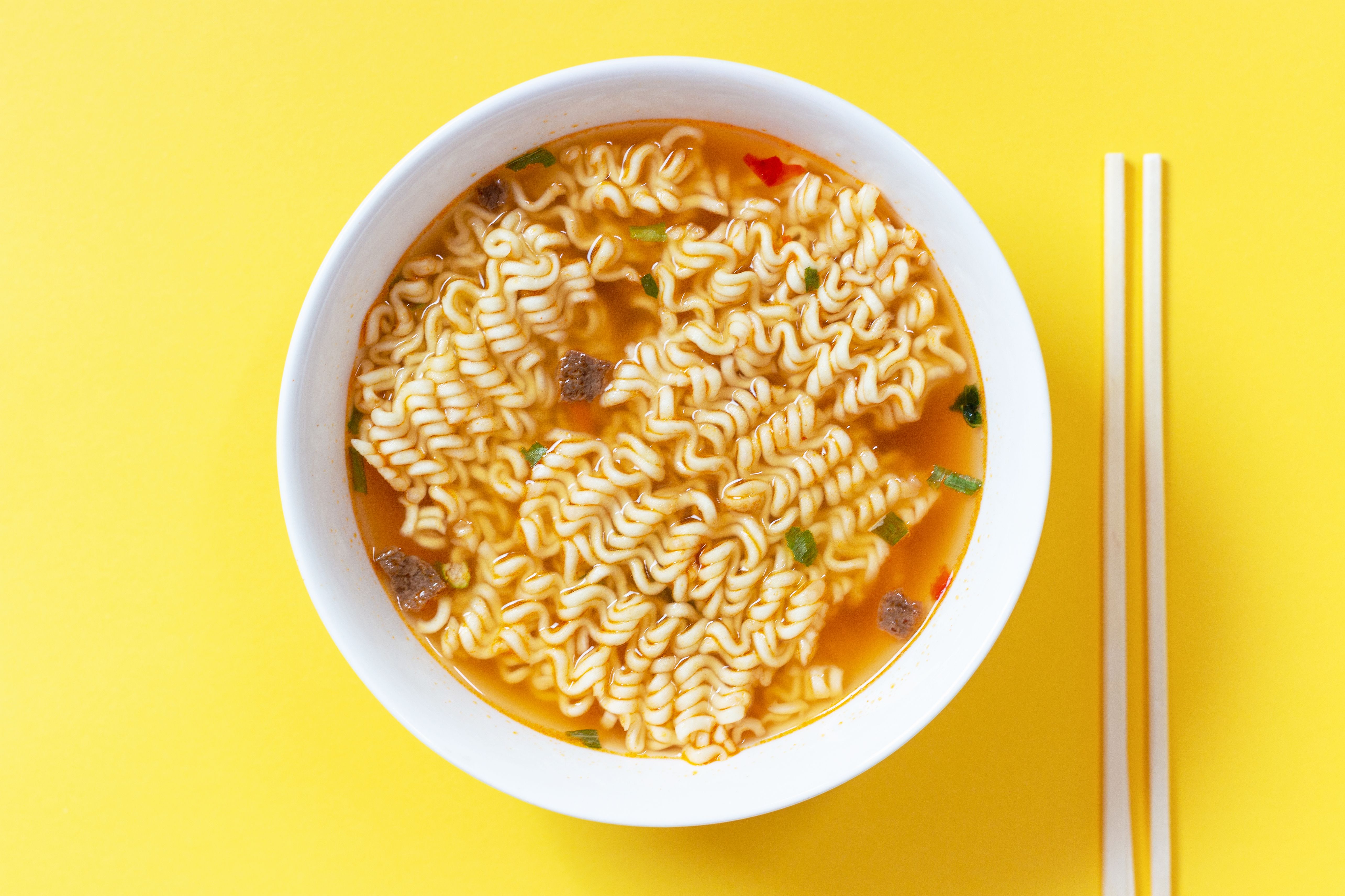 Instant-Nudelsuppe in einer weißen Schüssel auf gelbem Hintergrund, daneben Essstäbchen.