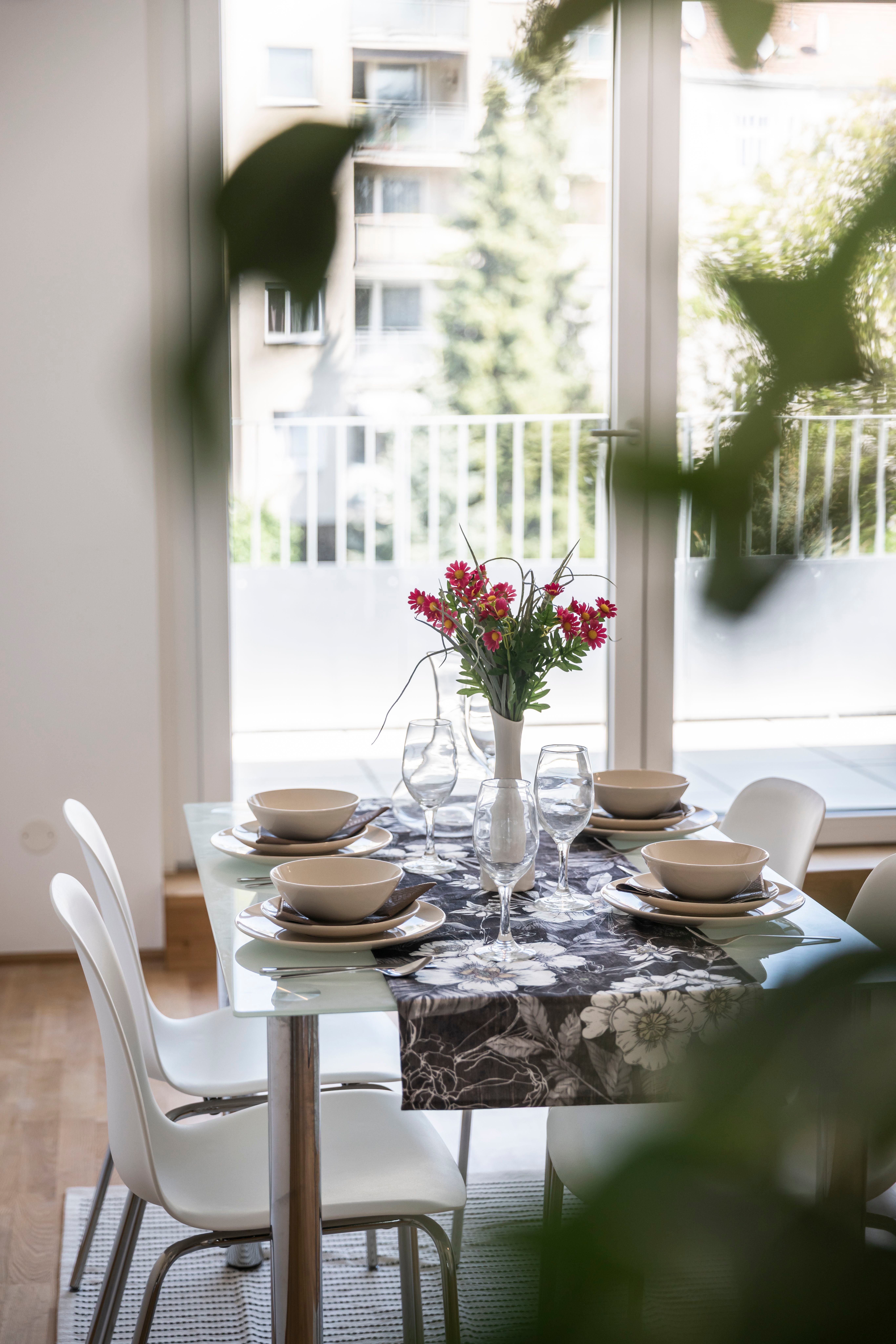 Modern gedeckter Esstisch mit Blumenstrauß vor großen Fenstern und Stühlen.