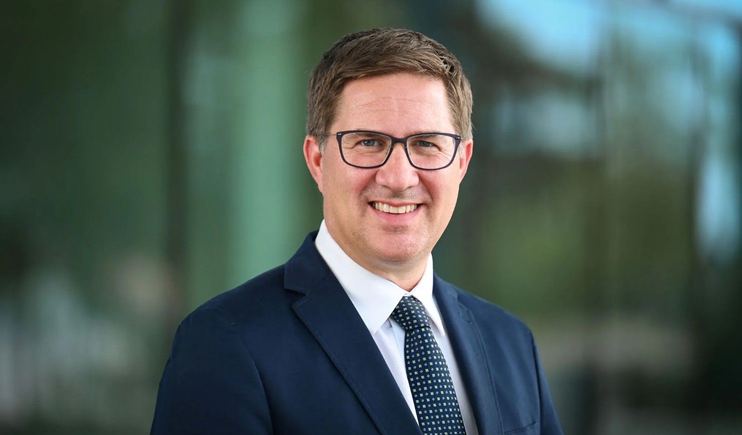 Ein Mann im Anzug und mit Brille lächelt in die Kamera, unscharfer Hintergrund.