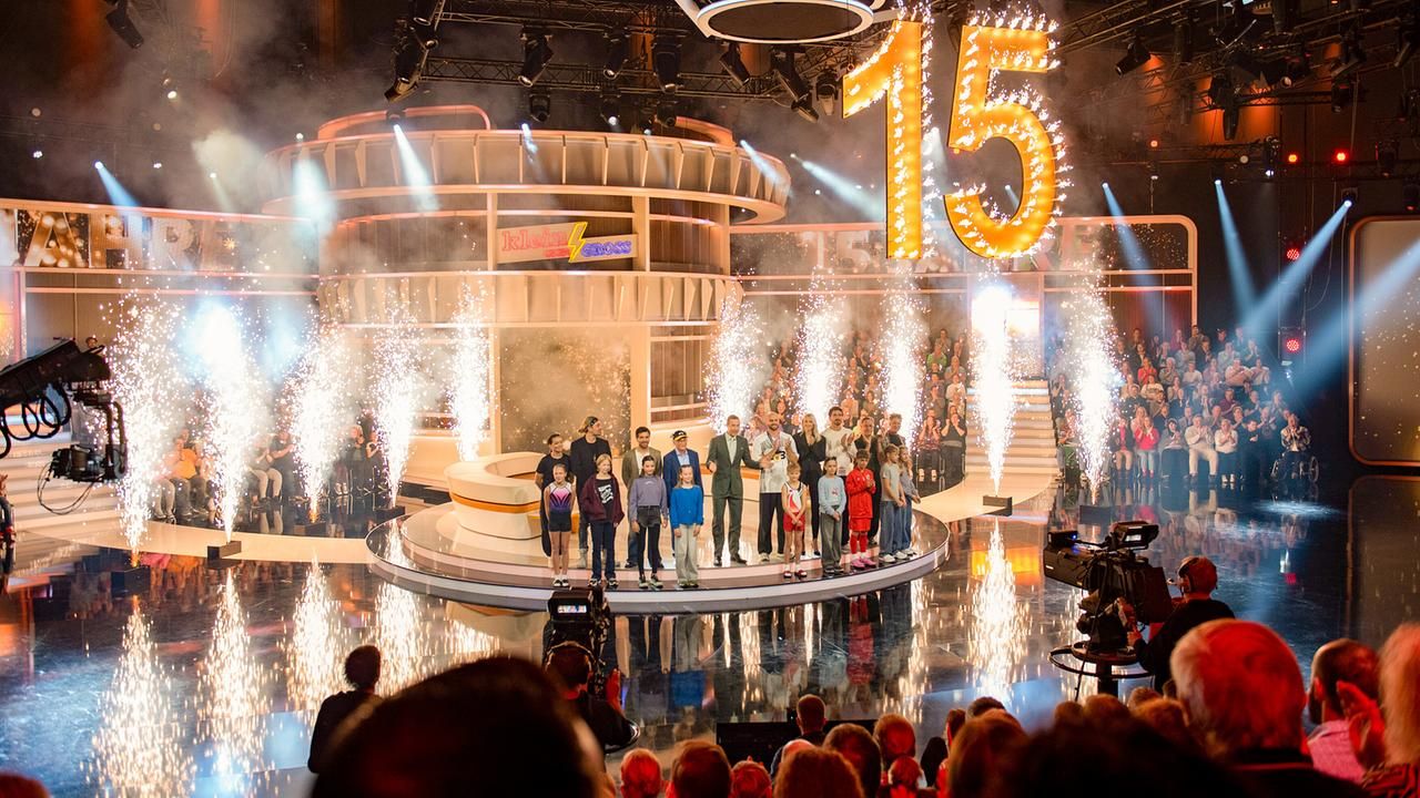 Bühnenbild einer Fernsehshow mit Feuerwerk und großer leuchtender 15, Menschen stehen im Rampenlicht.