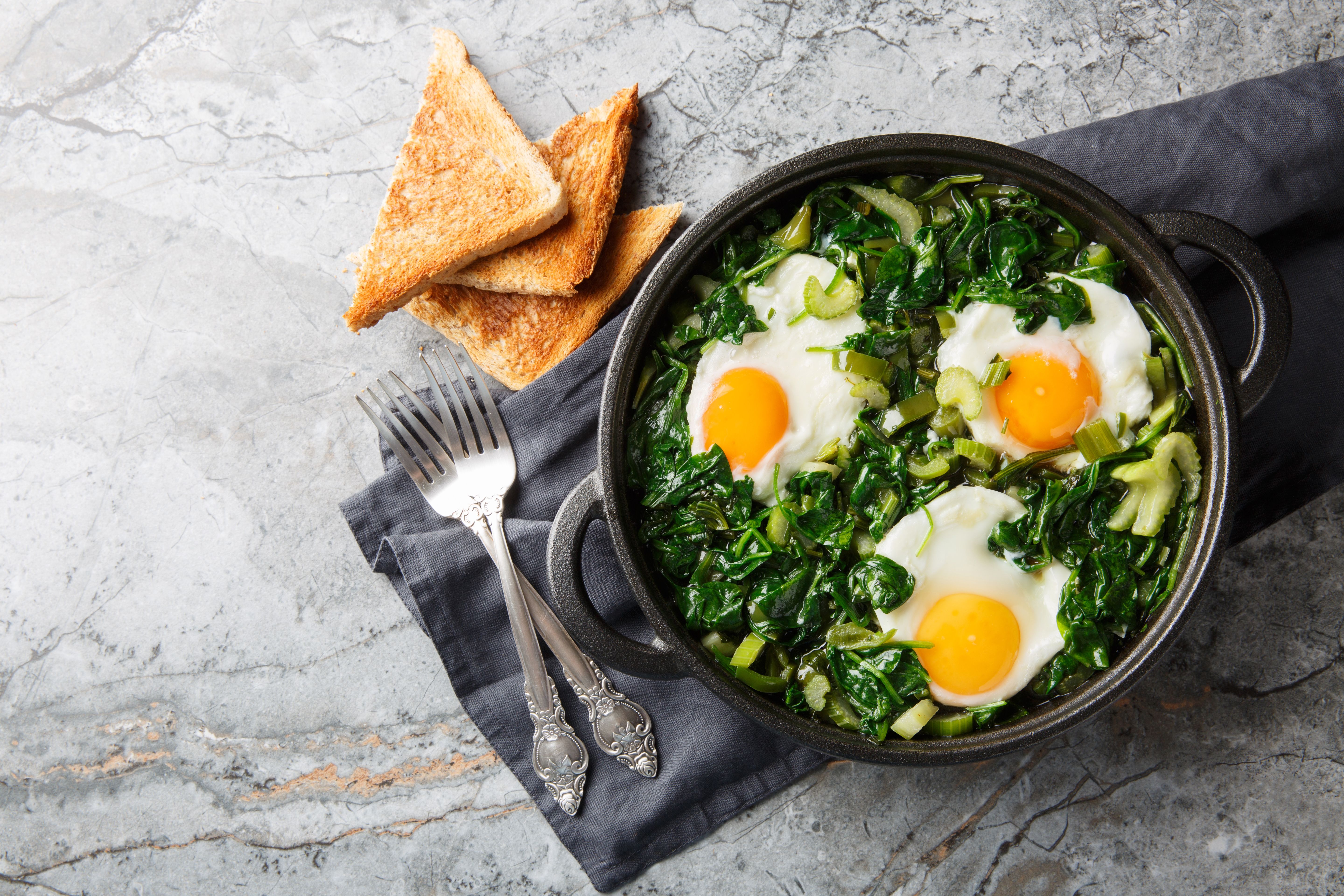 Grüne Shakshuka mit Spinat, Kräutern und drei Spiegeleiern in einer Pfanne, daneben Toast.