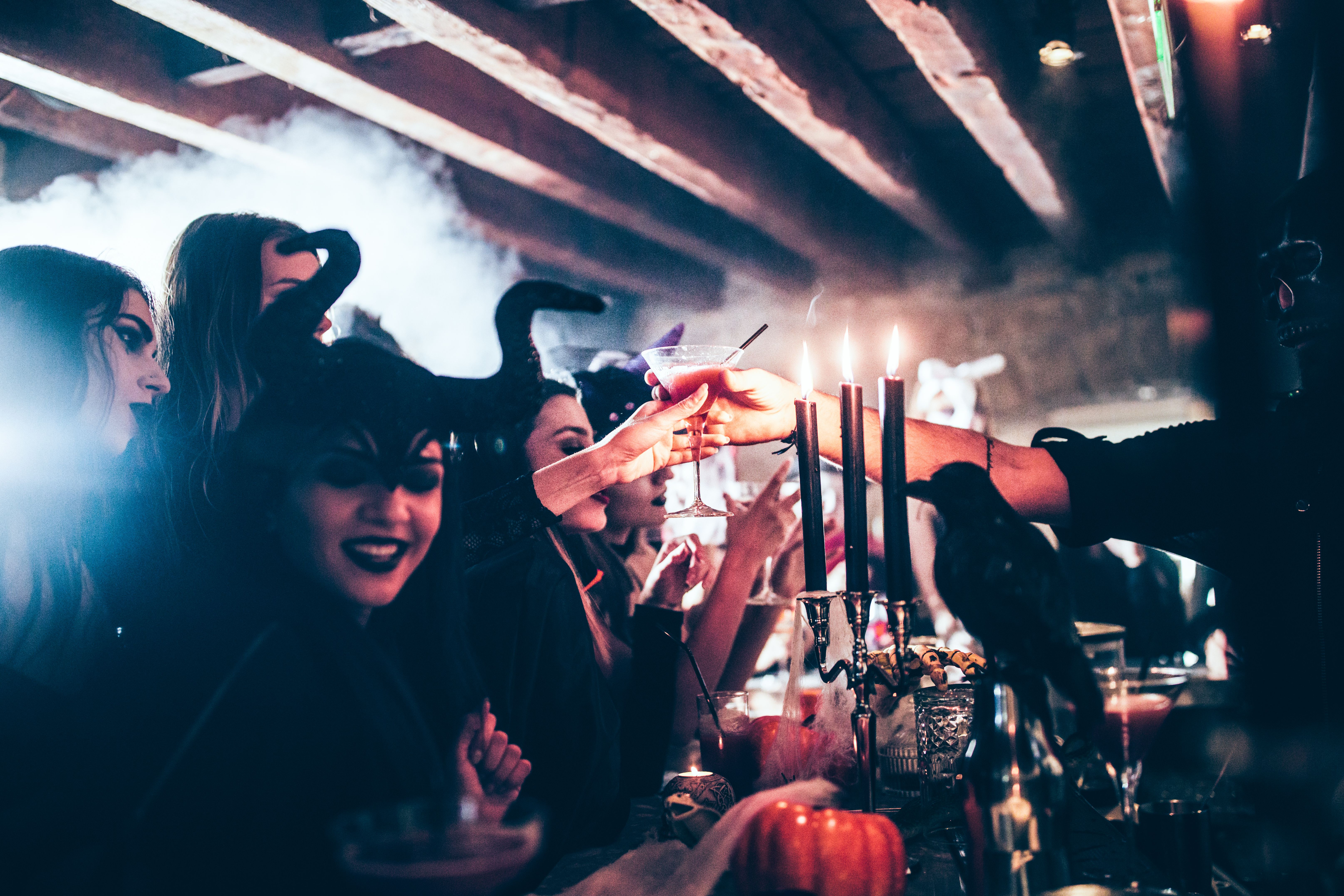 Barkeeper reicht einer Frau in Halloweenkostüm einen Cocktail, Kerzen leuchten auf dem Tresen.