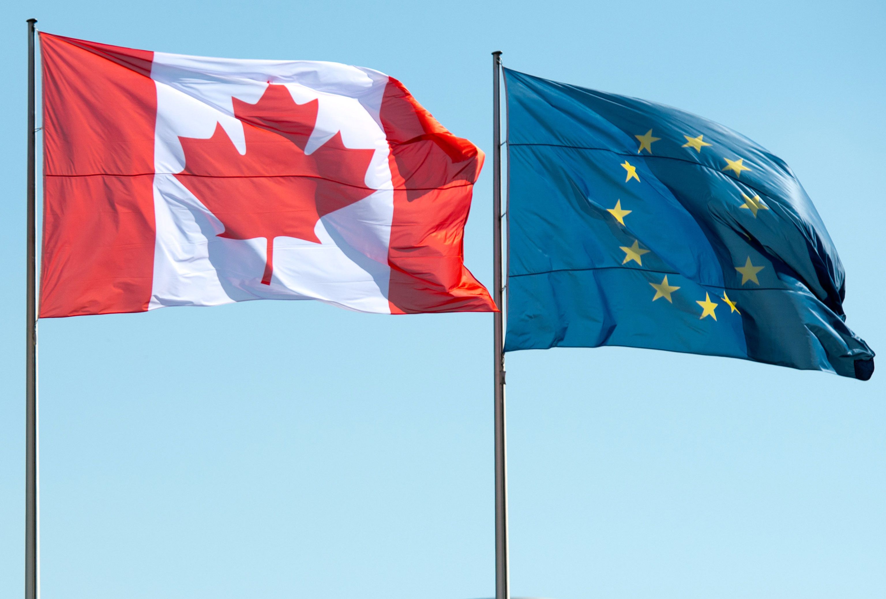 Die Flaggen von Kanada und der Europäischen Union wehen nebeneinander im Wind vor blauem Himmel.
