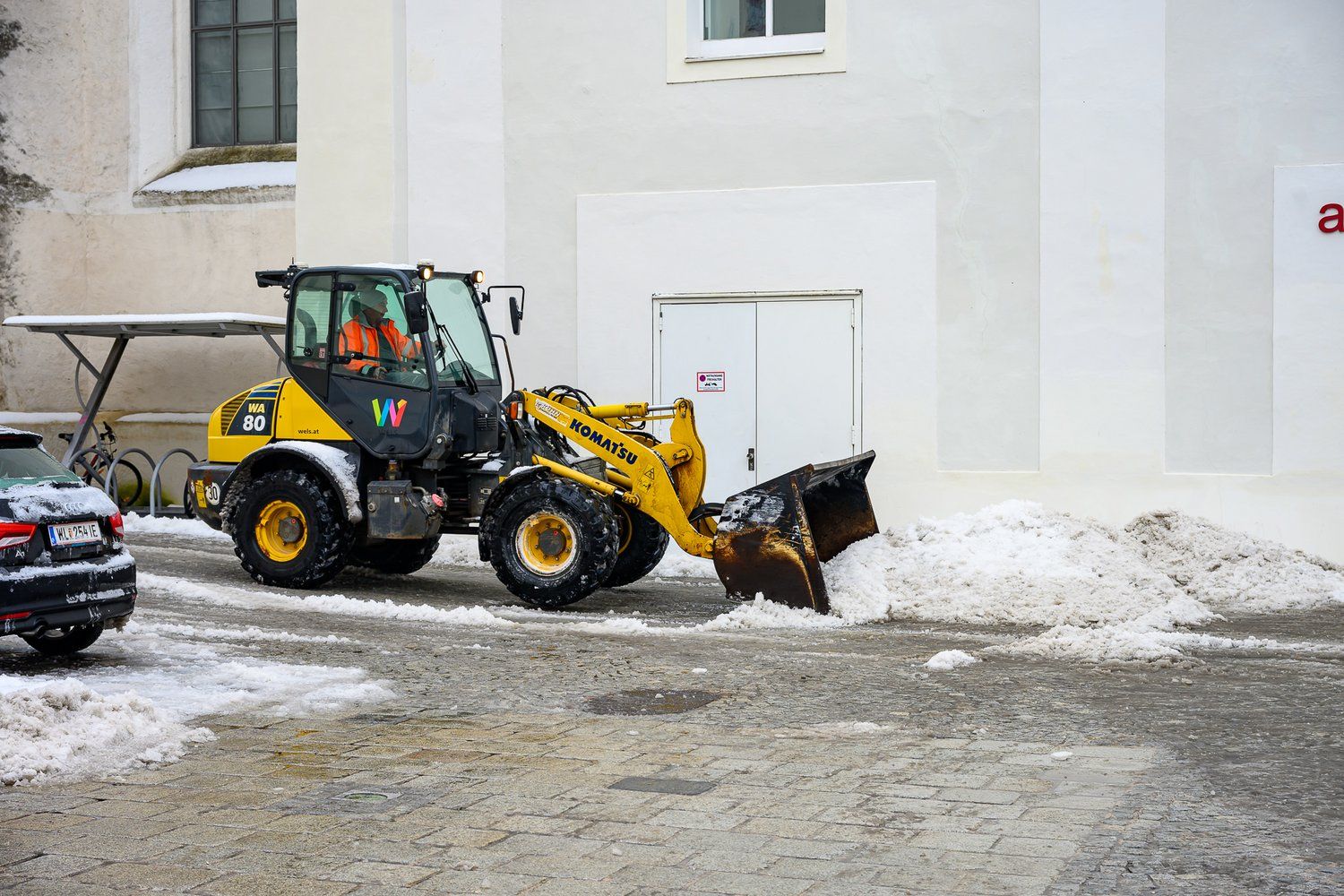 Ein Radlader räumt Schnee von einer gepflasterten Straße vor einem Gebäude in der Stadt.