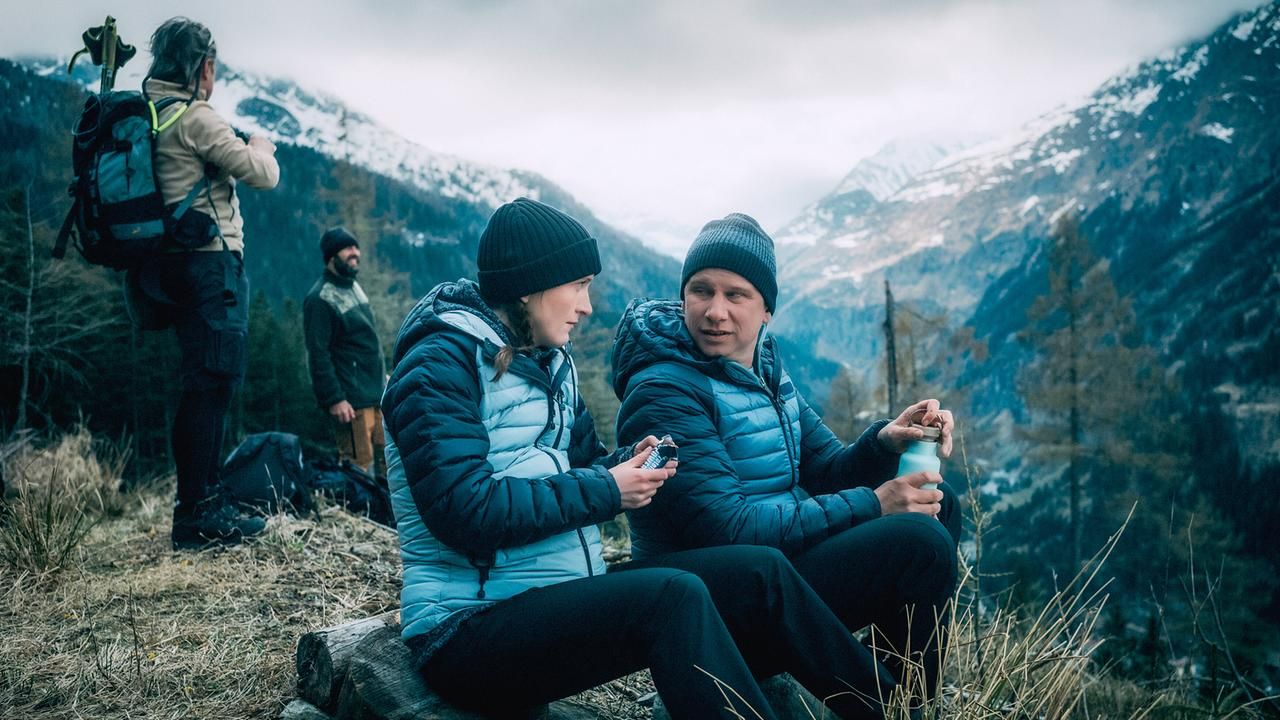 Vier Wanderer rasten in den Bergen, zwei sitzen und essen, zwei stehen mit Rucksäcken im Hintergrund.