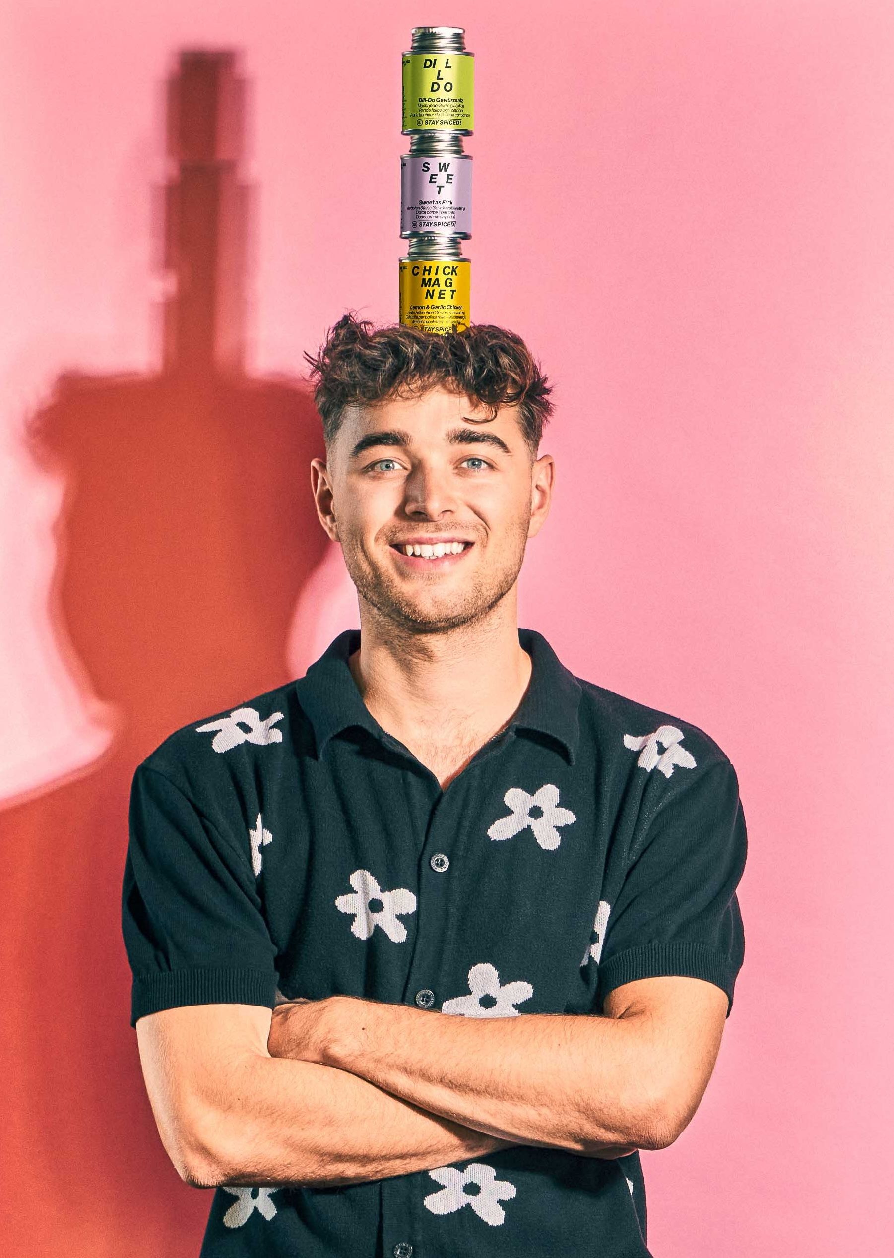 Junger Mann balanciert drei STAY SPICED!-Gewürzdosen auf seinem Kopf vor rosa Hintergrund.