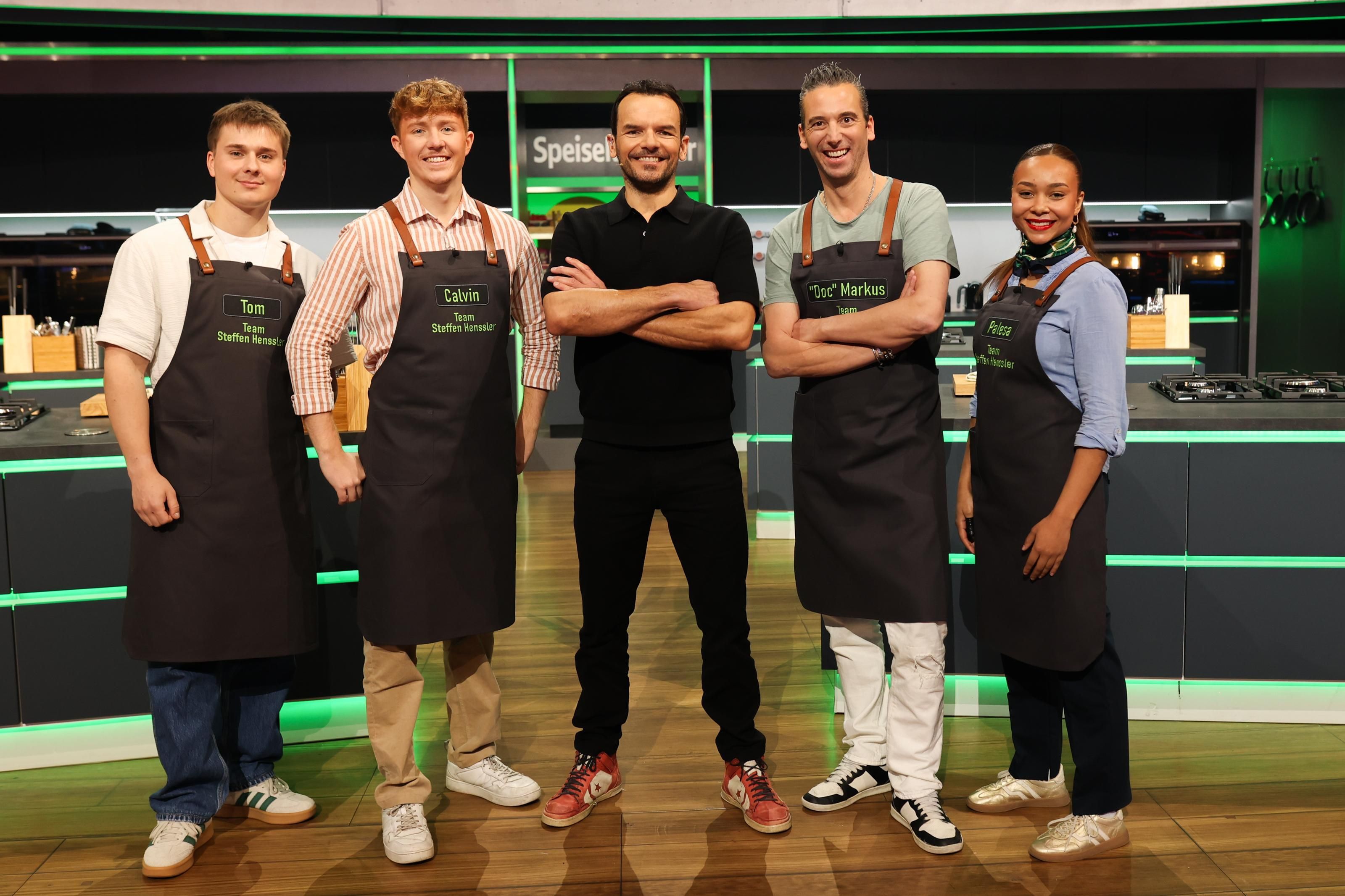 Fünf Personen stehen in einer Kochshow-Küche nebeneinander und lächeln in die Kamera.
