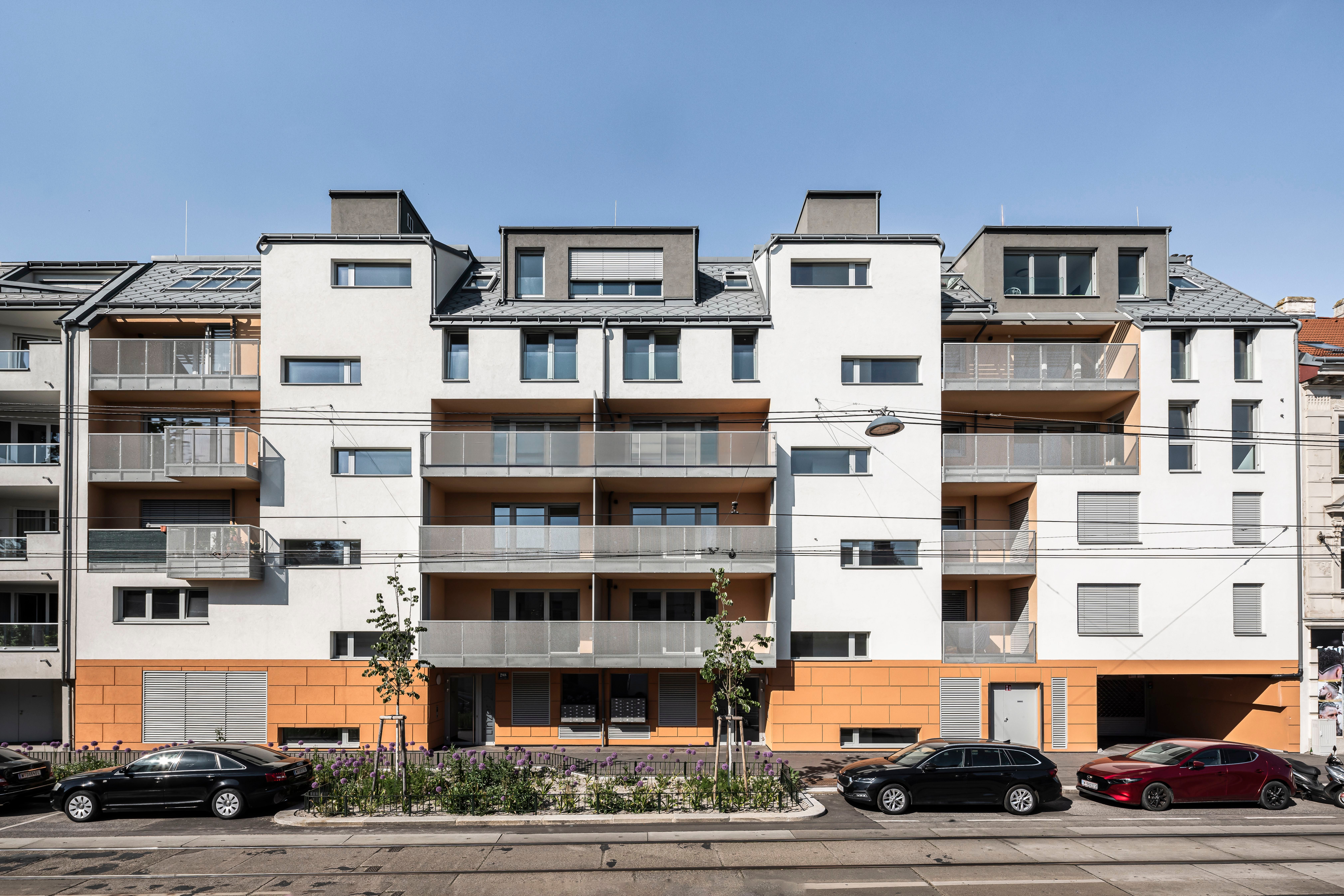 Modernes Wohngebäude mit Balkonen, davor parken mehrere Autos an der Straße.