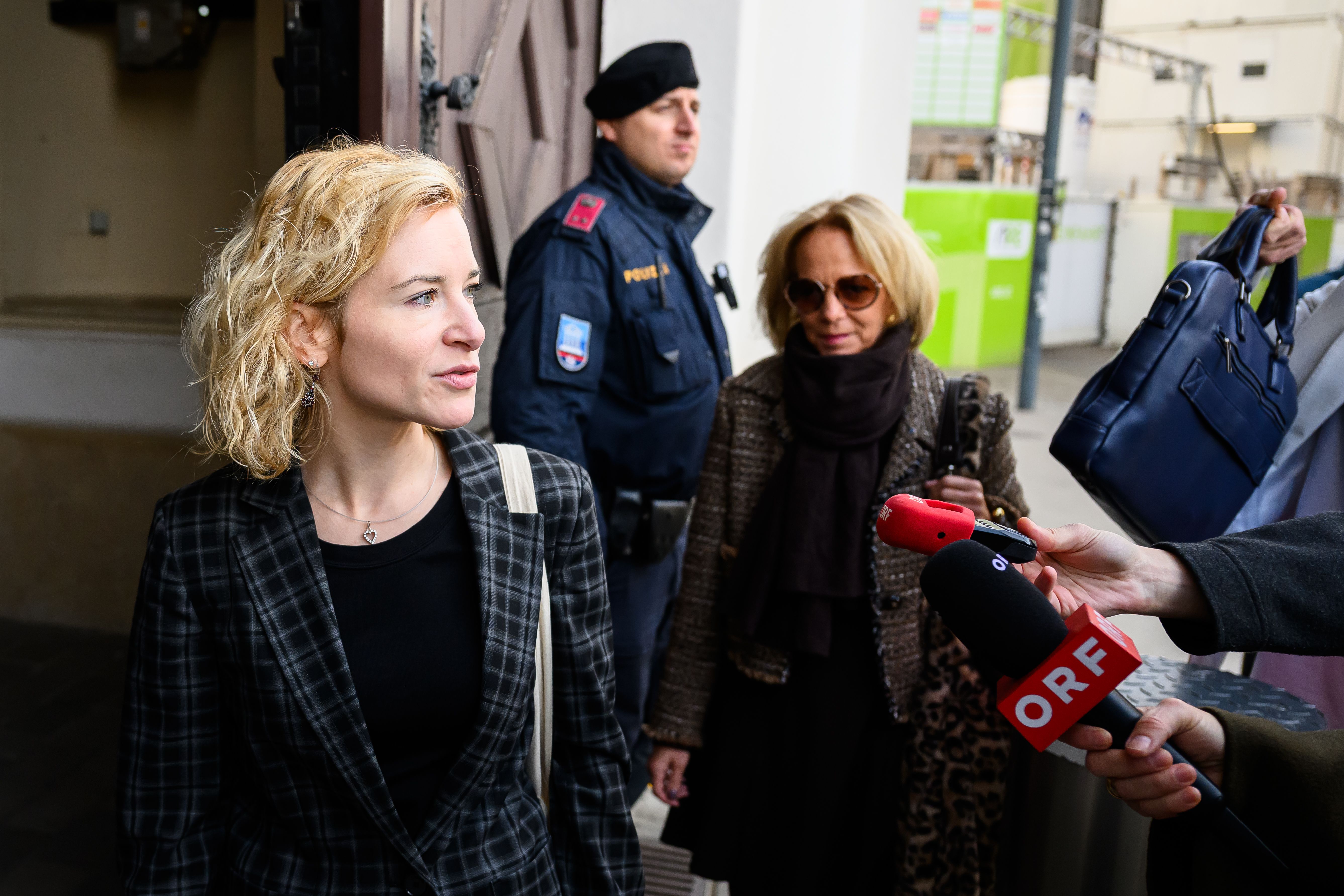 Eine Frau steht vor Mikrofonen, im Hintergrund Polizei und weitere Personen, draußen in Wien.
