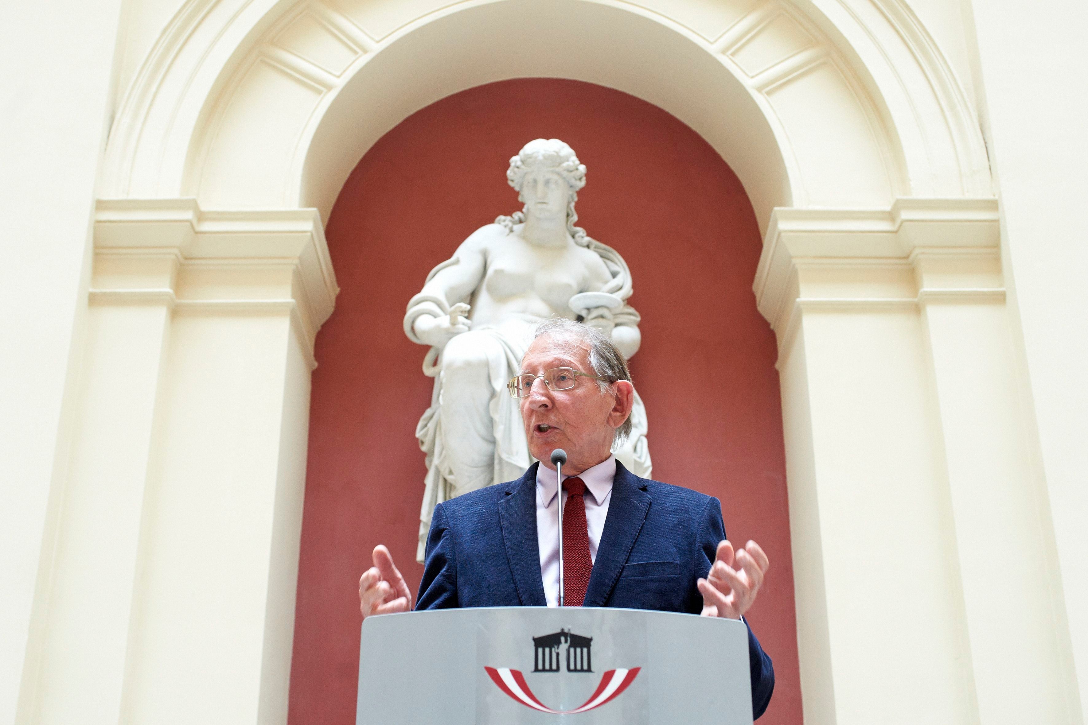 Ein Mann spricht am Rednerpult vor einer großen weißen Statue in einer Halle.