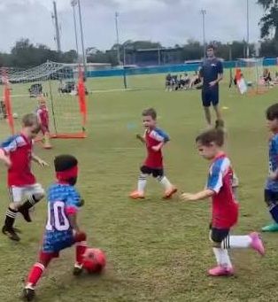 Mehrere Kinder spielen Fußball auf einem Rasenplatz, ein Erwachsener steht im Hintergrund.
