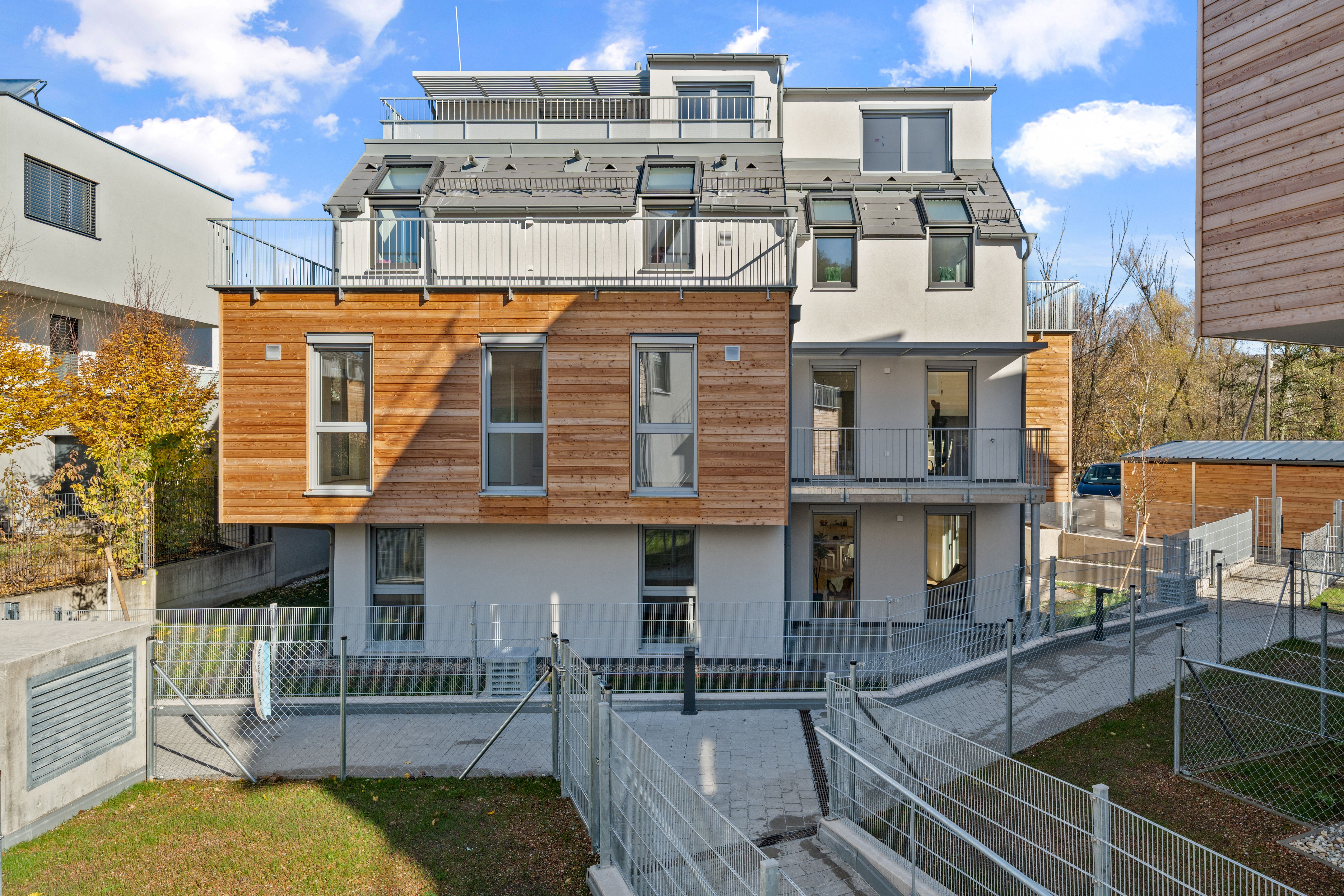 Modernes Mehrfamilienhaus mit Holz- und Putzfassade, großen Fenstern und Balkonen, umgeben von Zäunen.