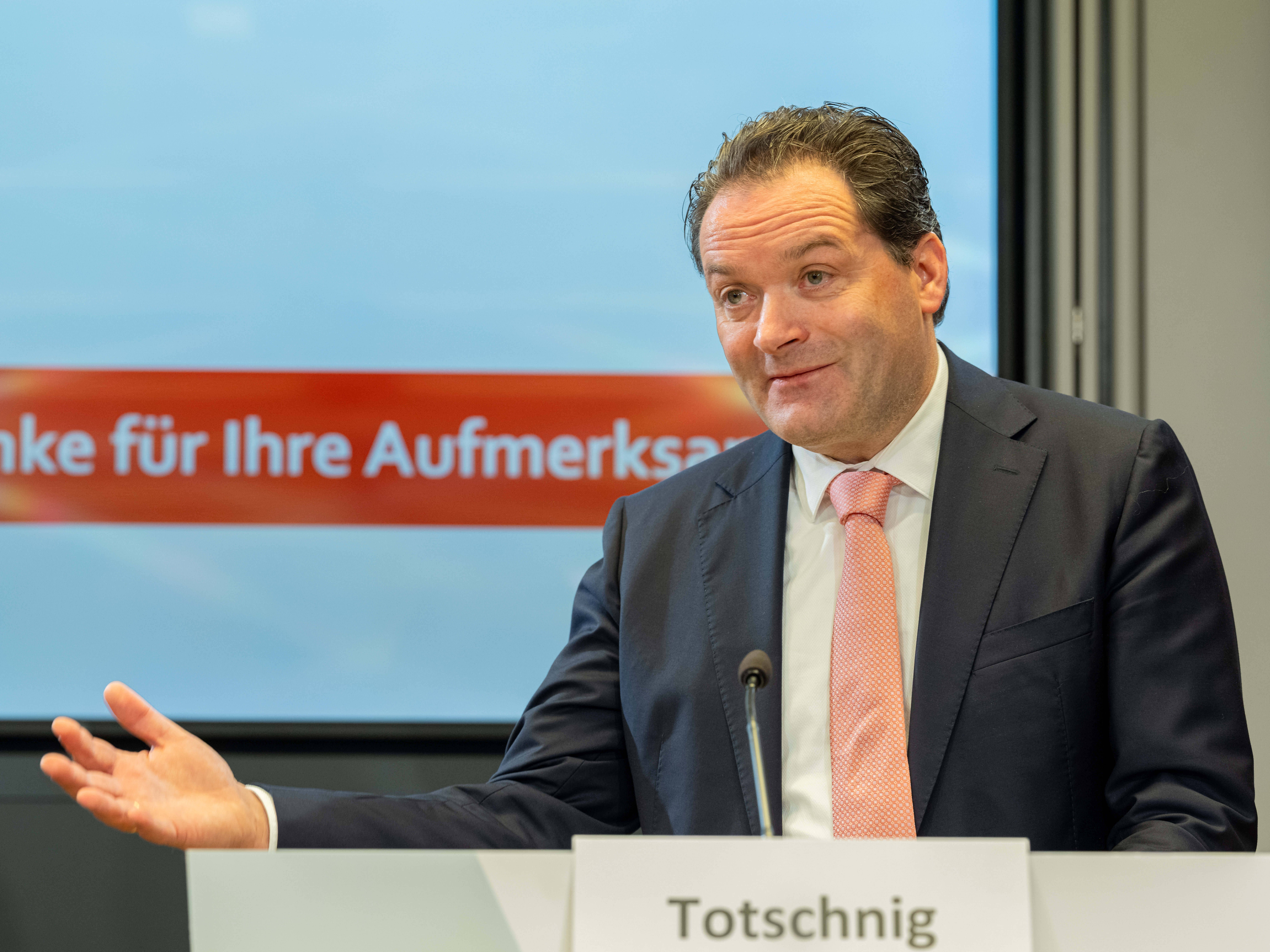 Ein Mann im Anzug spricht an einem Rednerpult mit dem Namensschild "Totschnig" bei einer Präsentation.