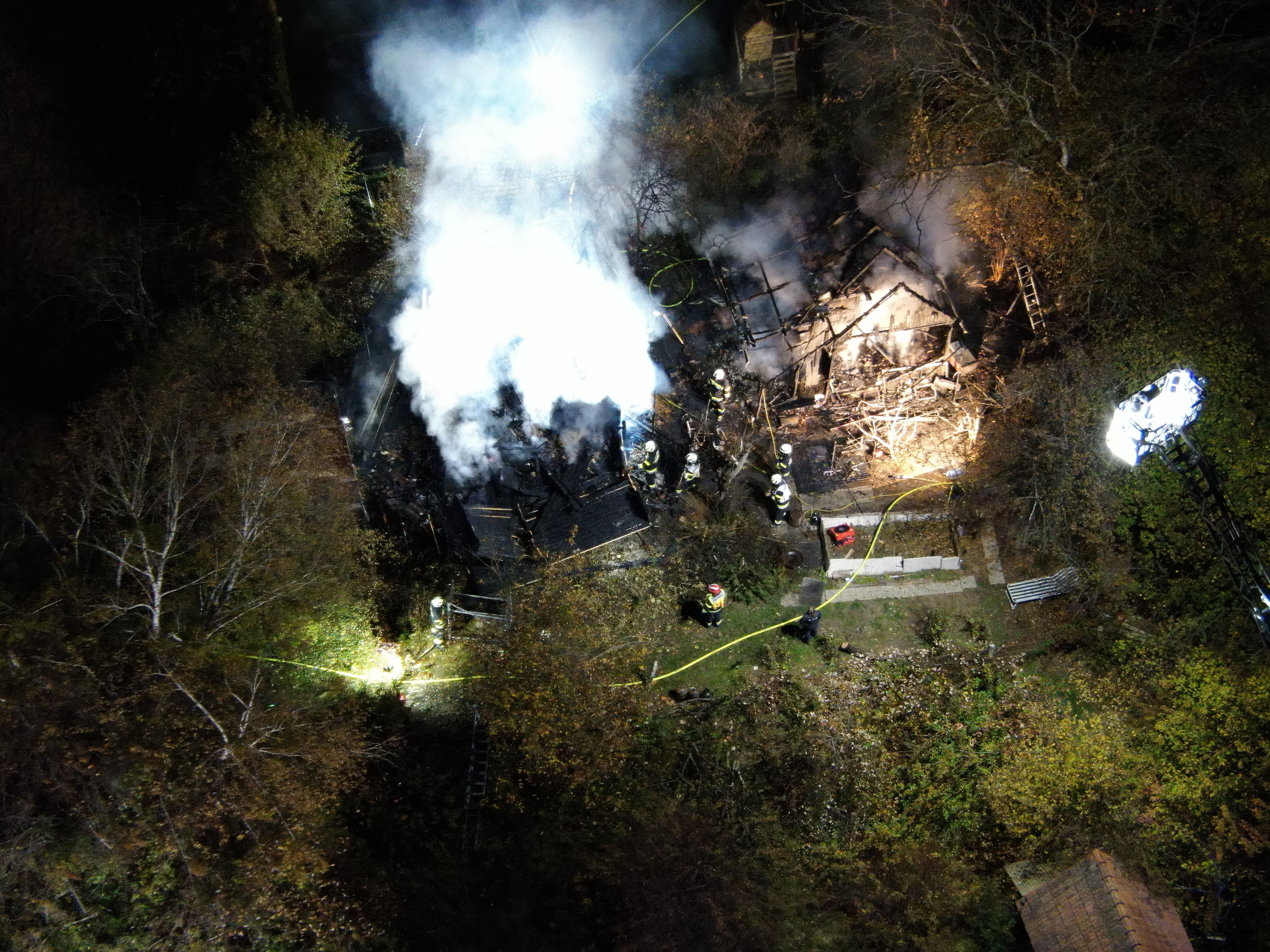 Feuerwehr löscht bei Nacht ein brennendes, stark qualmendes Holzhaus im Wald aus der Vogelperspektive.