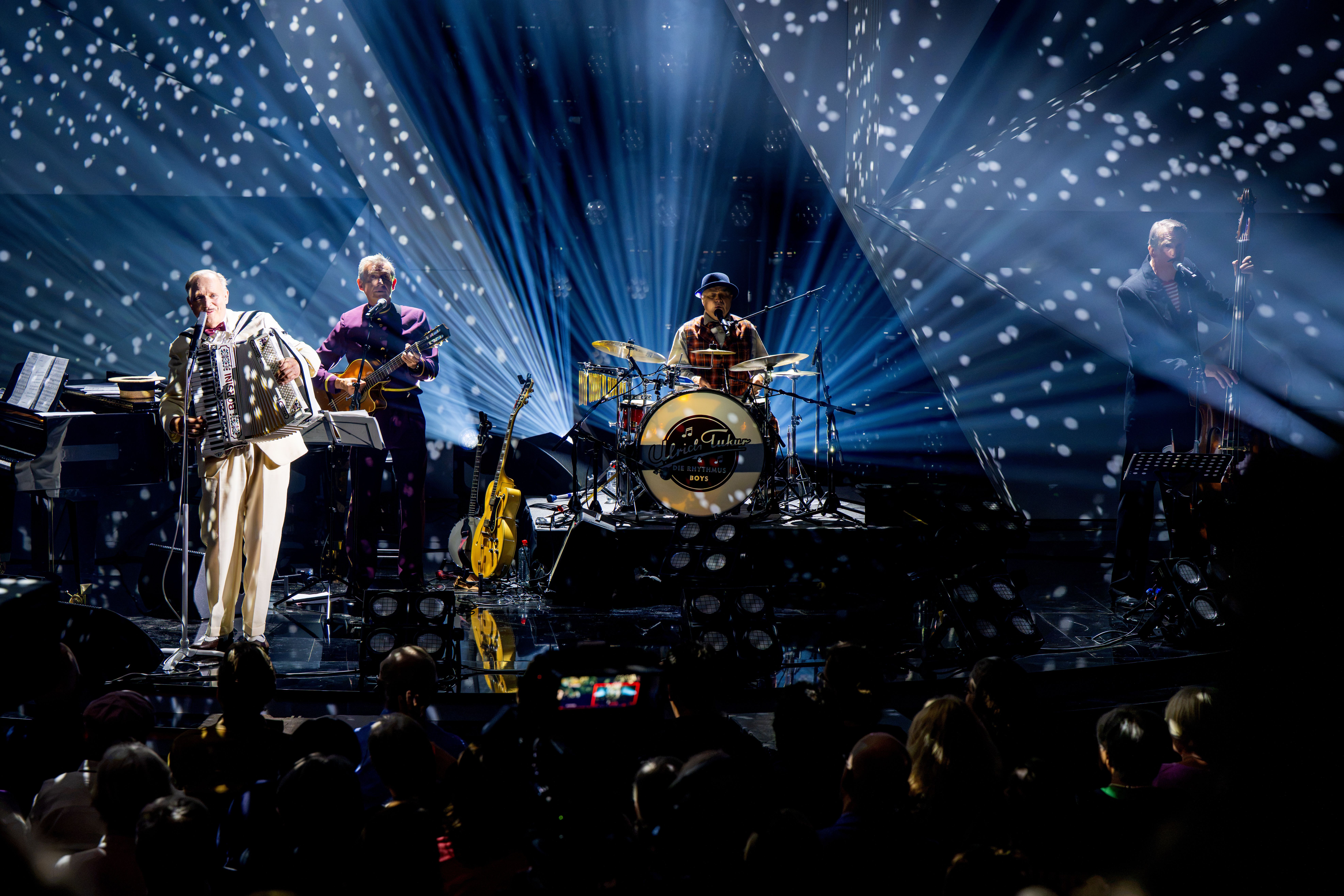 Ulrich Tukur & Die Rhythmus Boys spielen live auf einer Bühne mit Lichteffekten und Publikum.