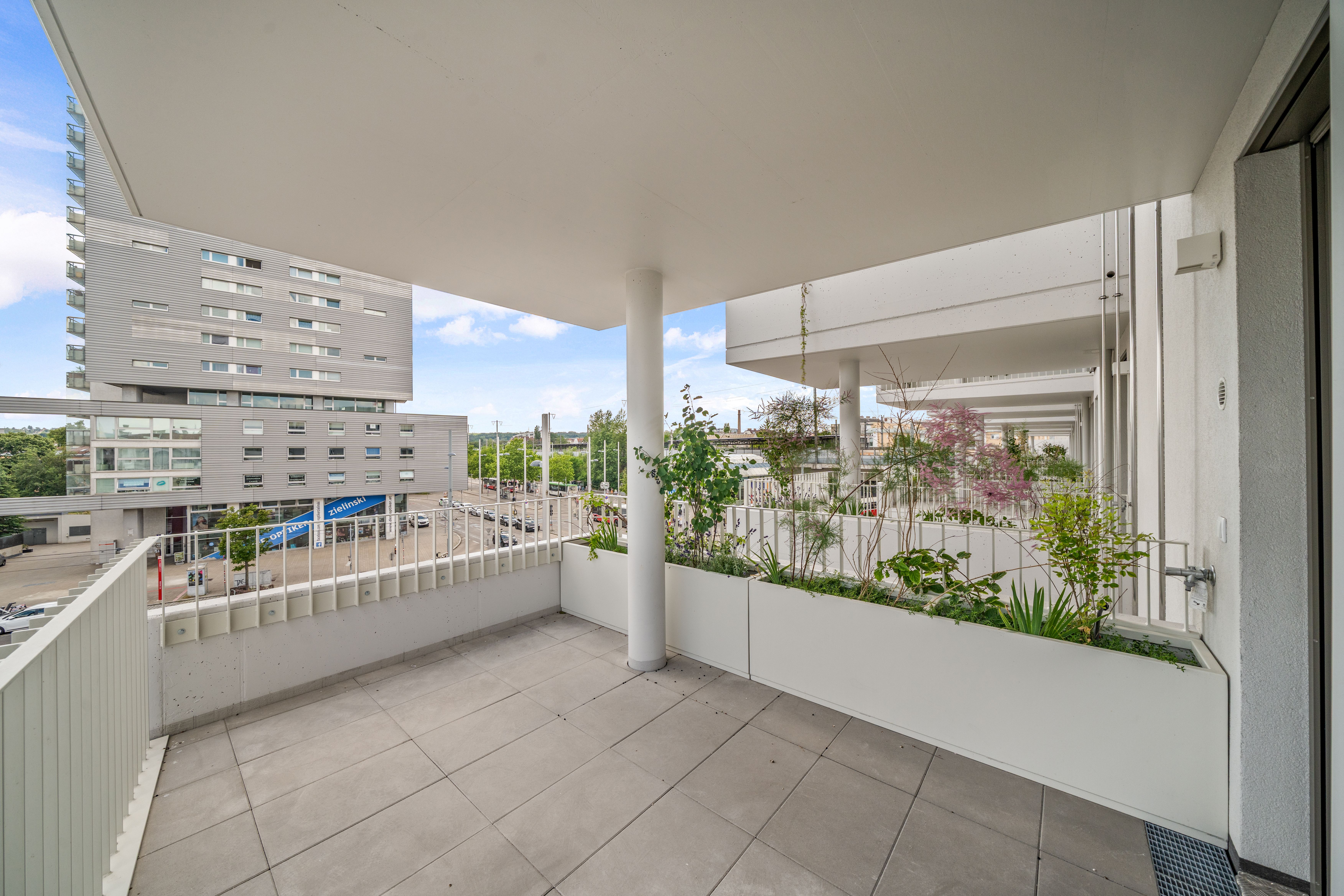 Großer, überdachter Balkon mit Pflanzkübeln und Blick auf moderne Stadtgebäude.