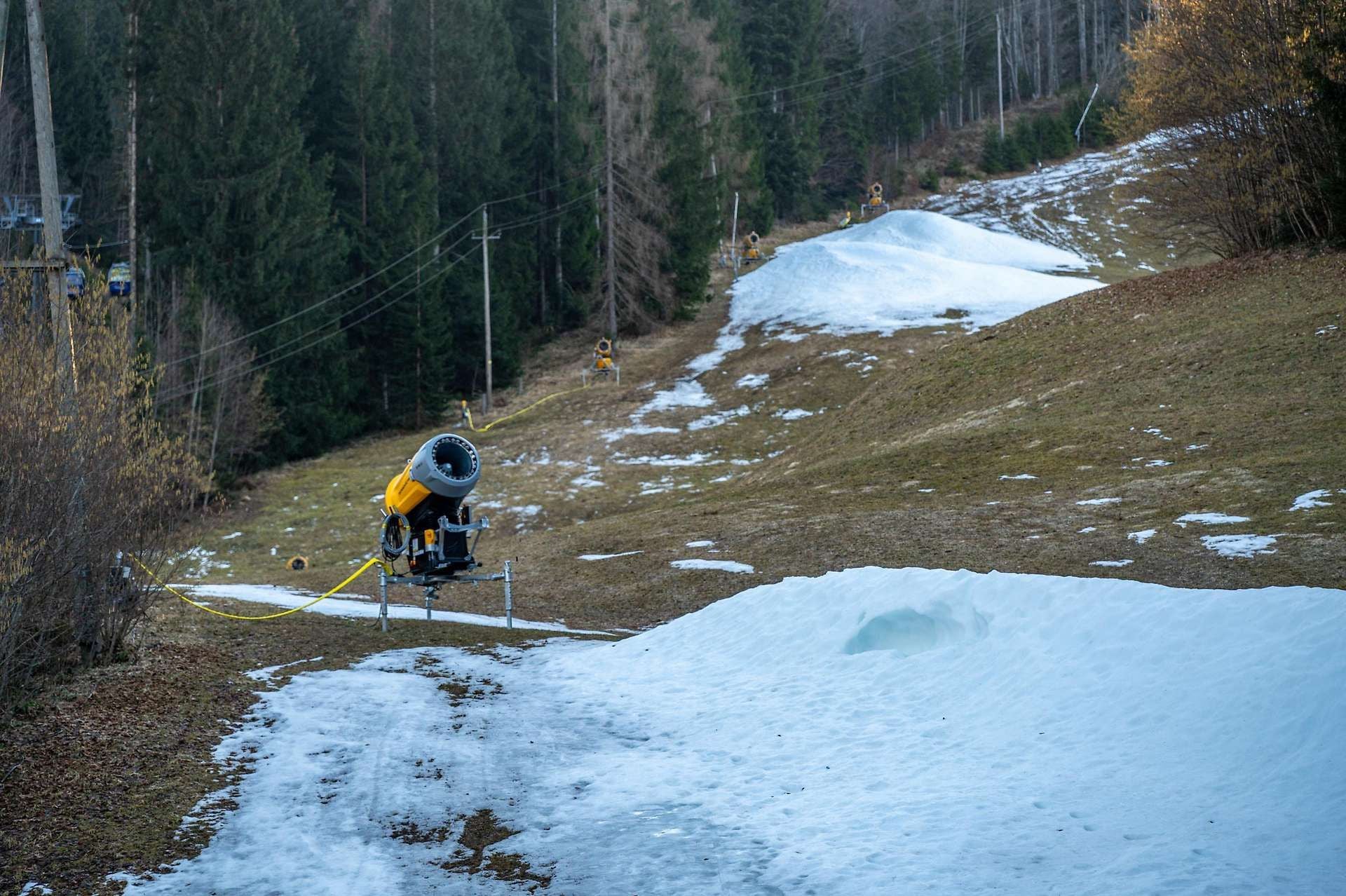 Eine Schneekanone steht auf einer teilweise schneebedeckten Piste in hügeliger Landschaft.
