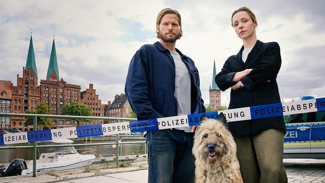 Zwei Personen und ein Hund stehen vor einer Polizeiabsperrung am Wasser, im Hintergrund Altstadthäuser.