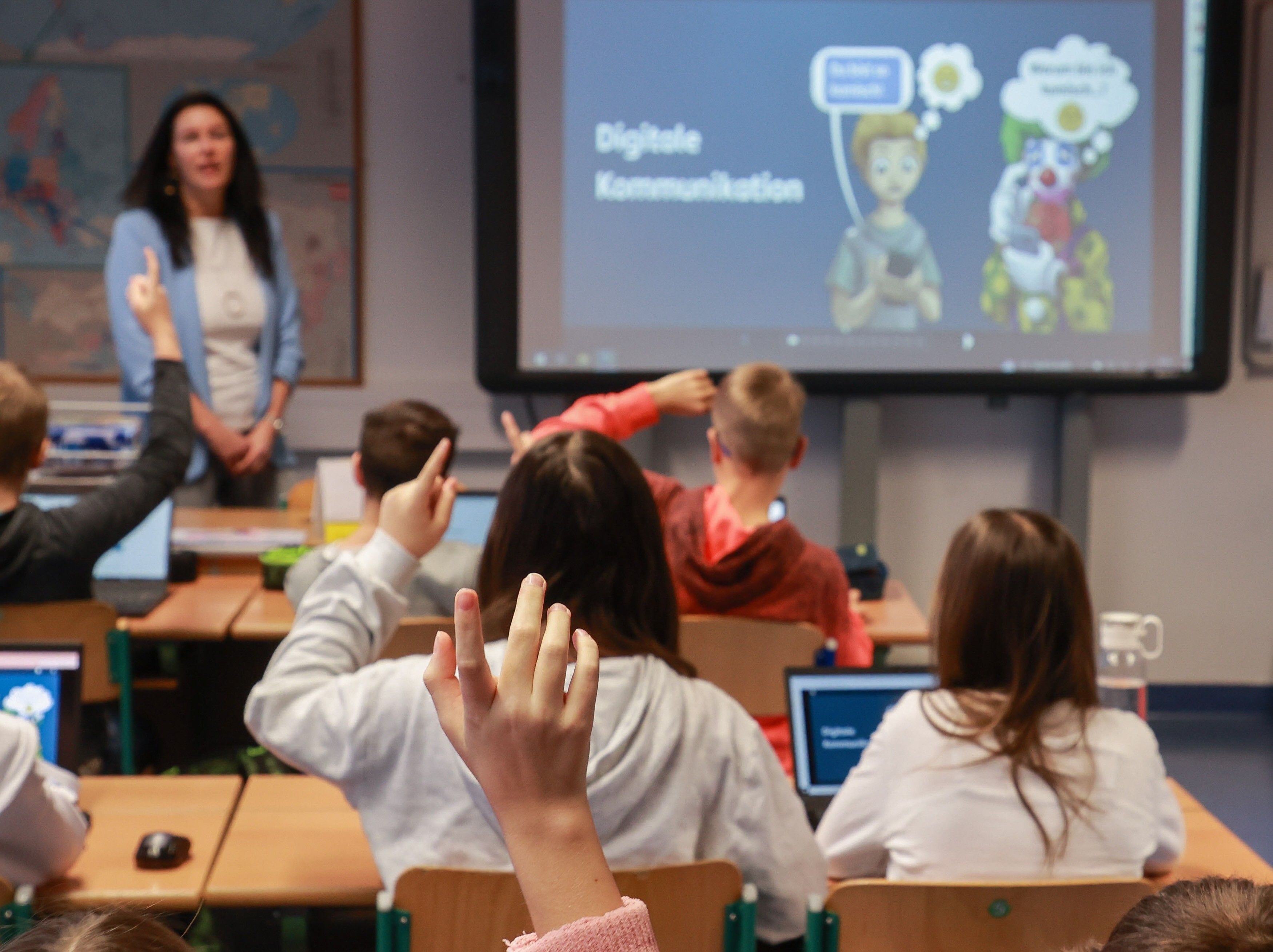 Schüler heben die Hand im Klassenraum; Lehrerin steht vorne, Tafel zeigt "Digitale Kommunikation".