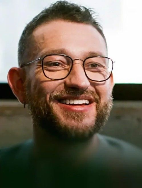 Ein lachender Mann mit Brille und Bart vor unscharfem Hintergrund.
