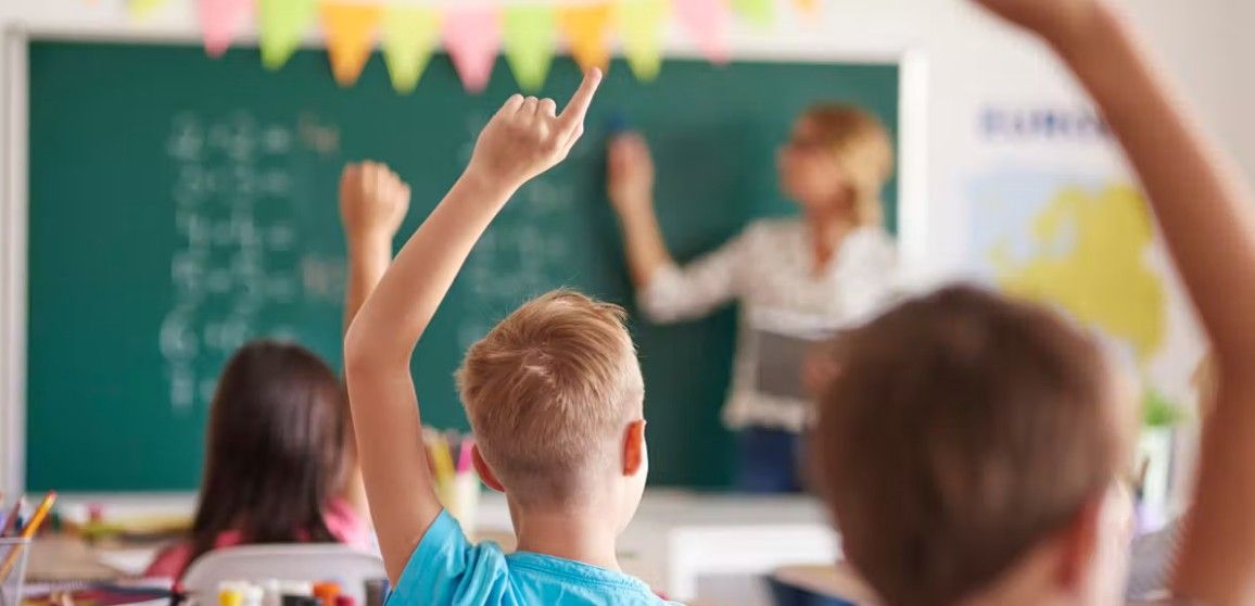 Kinder heben im Klassenzimmer die Hände, während eine Lehrerin an der Tafel unterrichtet.
