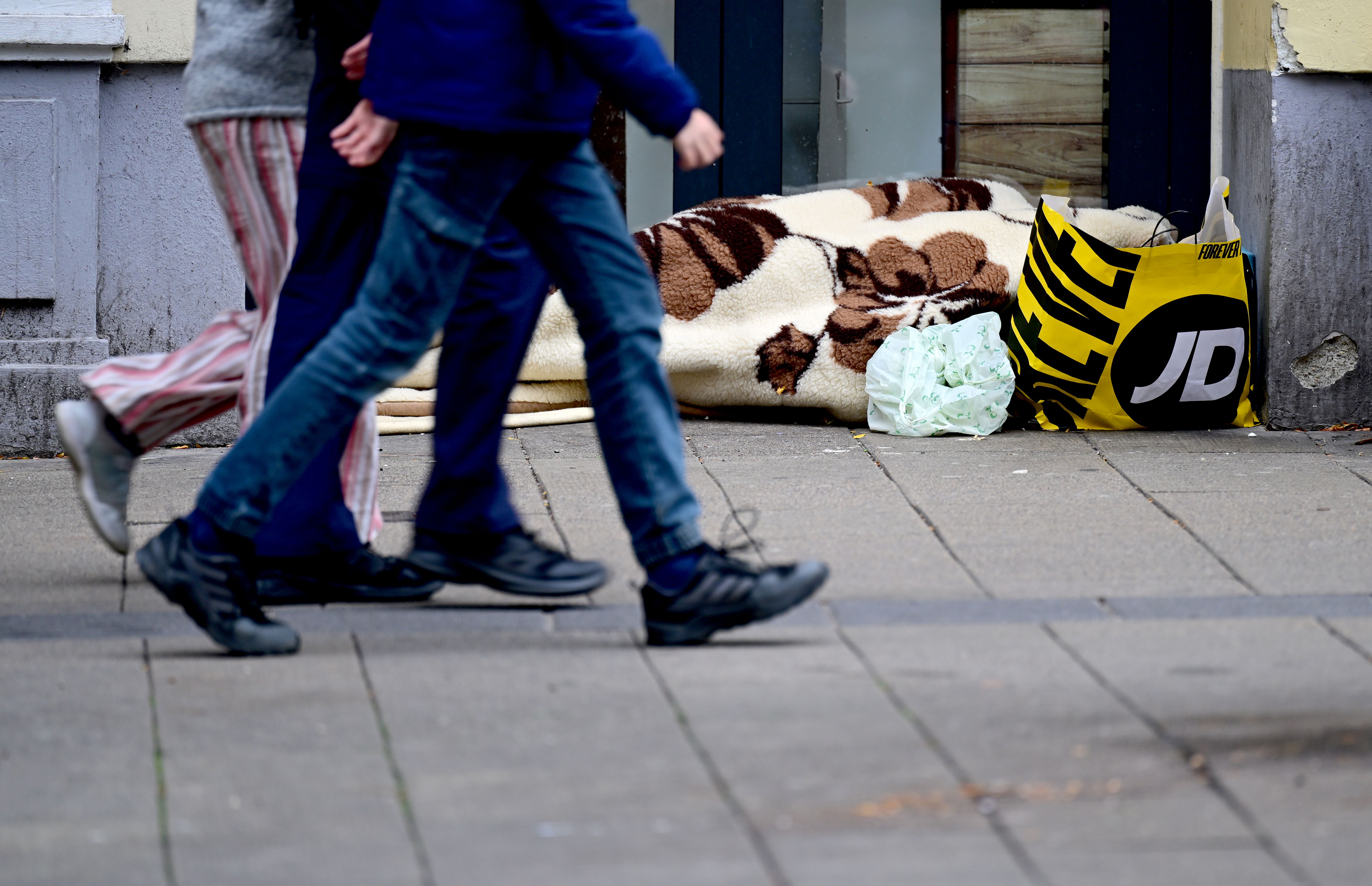 Eine obdachlose Person liegt in Decken gehüllt in einer Nische, Passanten gehen vorbei.