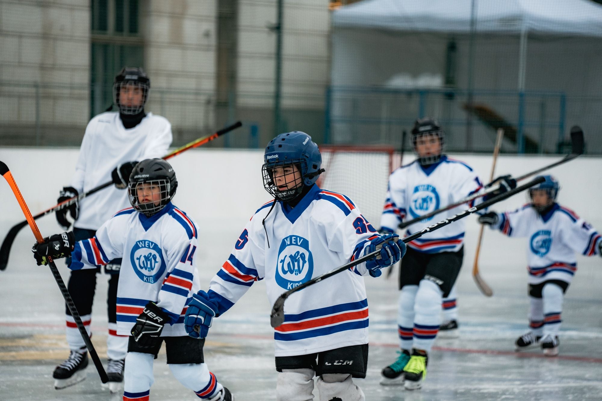 Mehrere Kinder spielen Eishockey auf einer Eisbahn, alle tragen WeV-Trikots und Helme.
