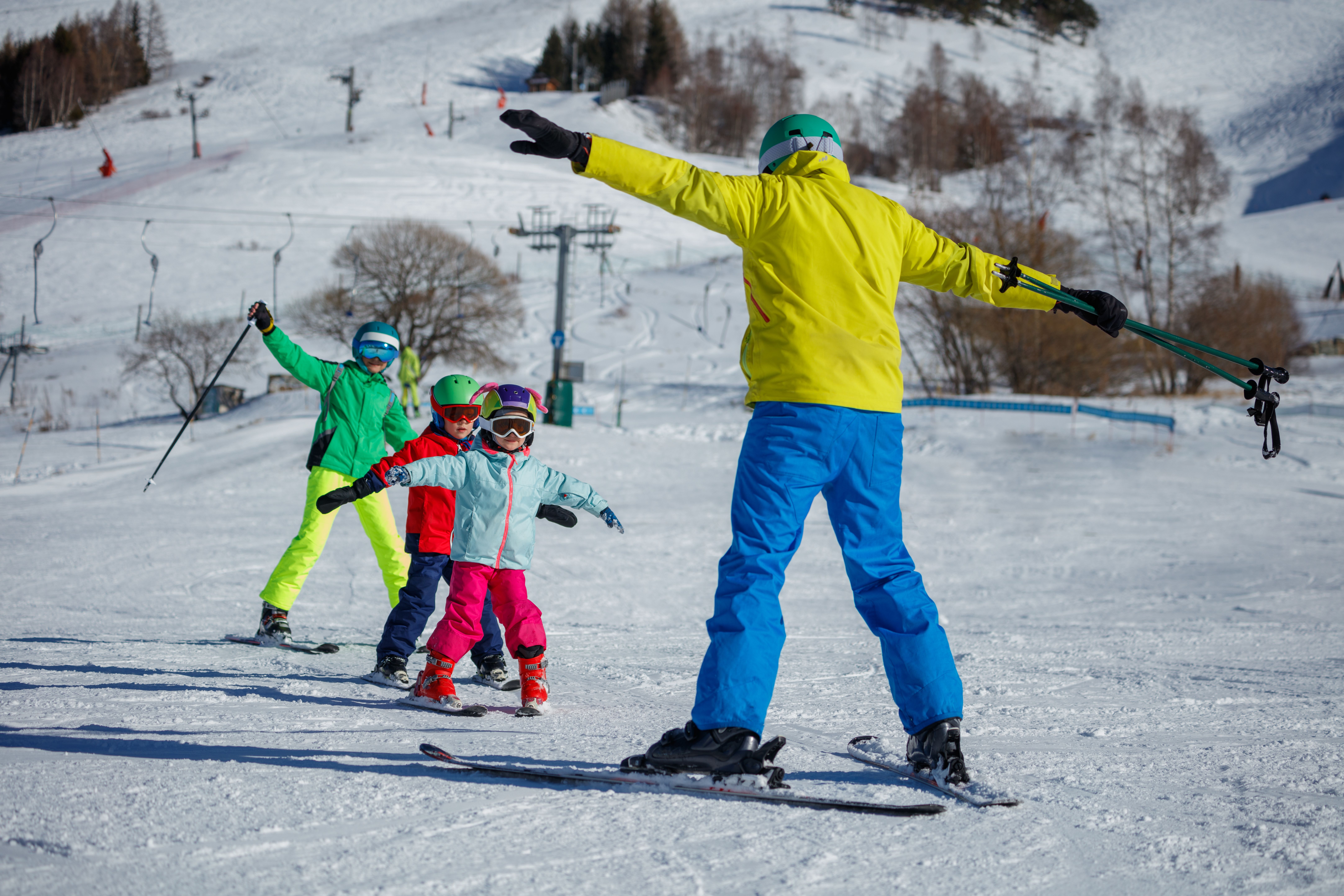 Skilehrer zeigt drei Kindern auf einer Skipiste Skitechniken, alle in bunter Winterkleidung.