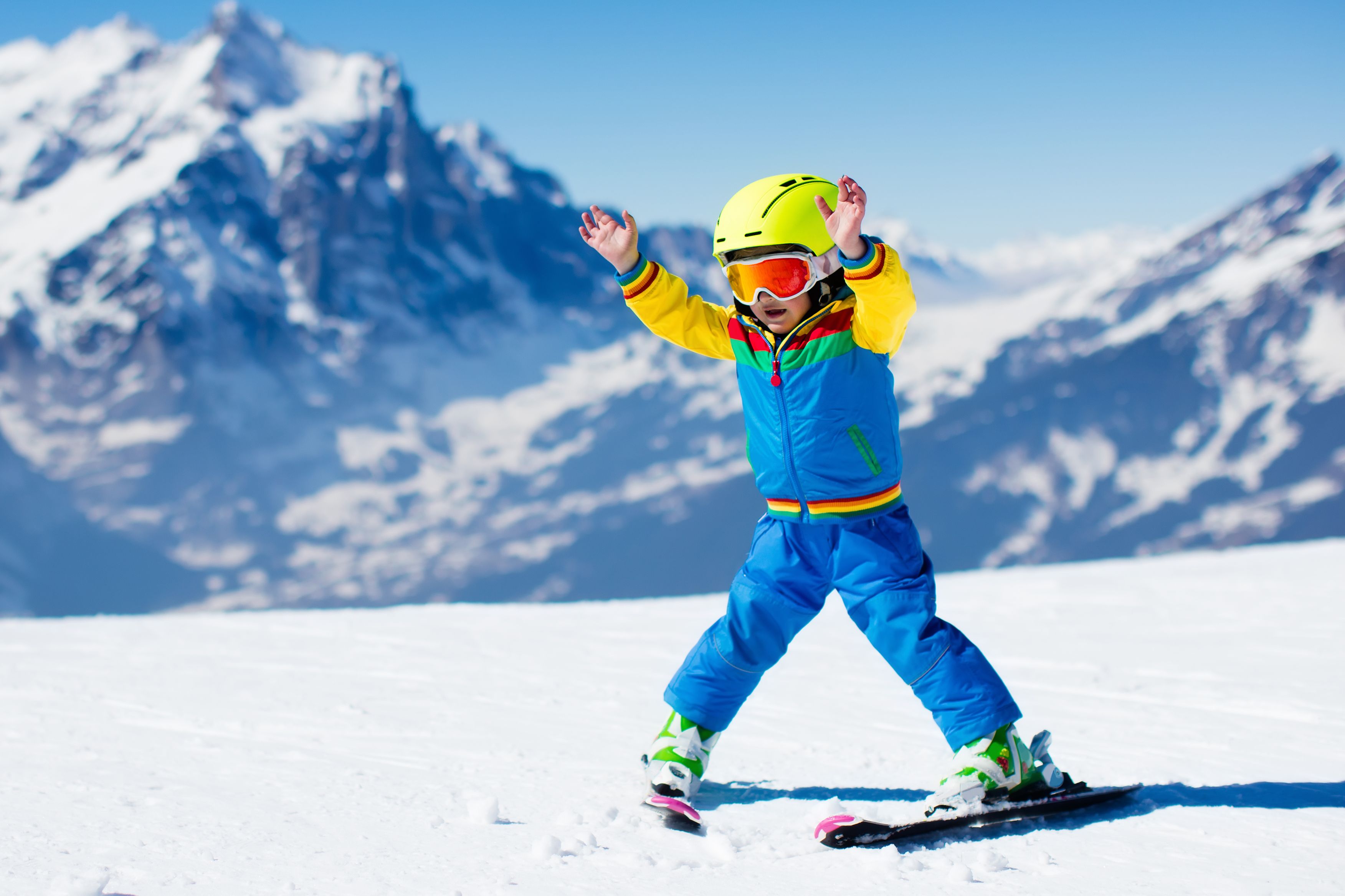 Kleines Kind mit Helm und Skibrille fährt Ski im Schnee vor verschneiten Bergen.