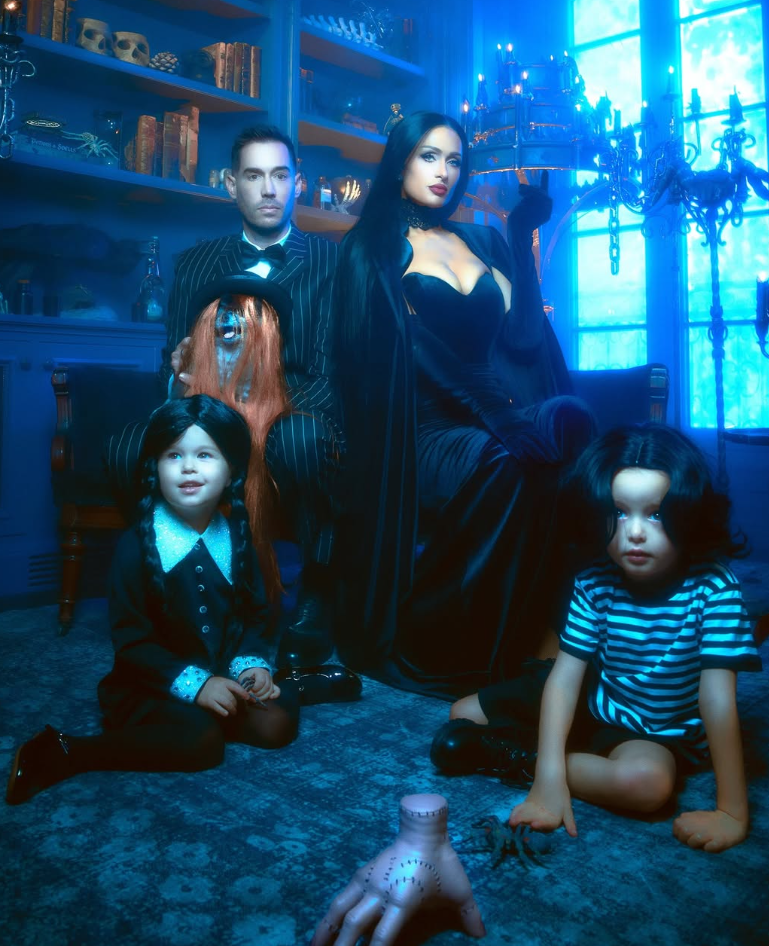 Familie im "Addams Family"-Kostüm posiert in einem düster dekorierten Raum mit blauer Beleuchtung.