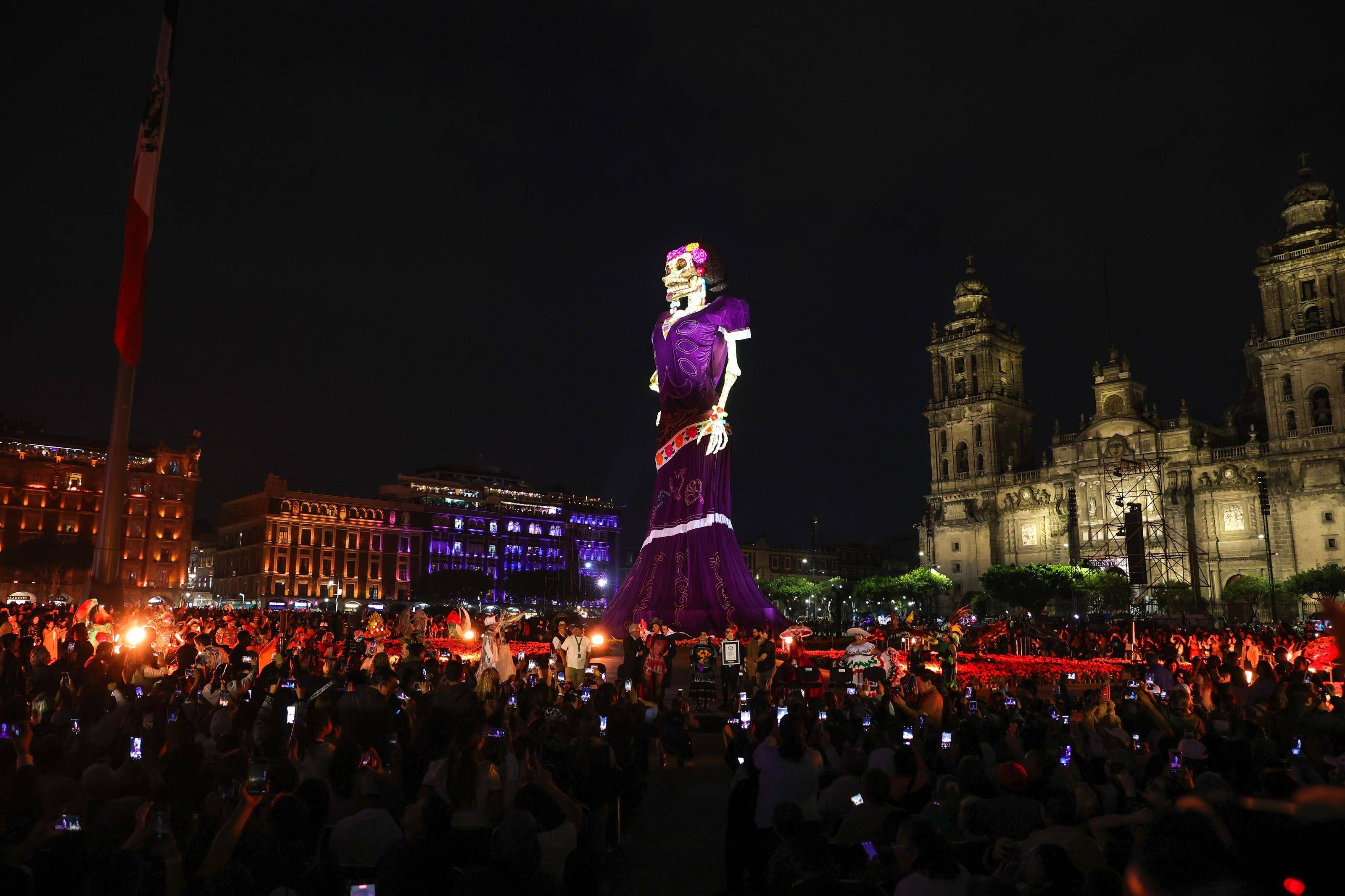 Menschenmenge feiert am Abend vor einer riesigen Catrina-Figur auf dem beleuchteten Zócalo-Platz.