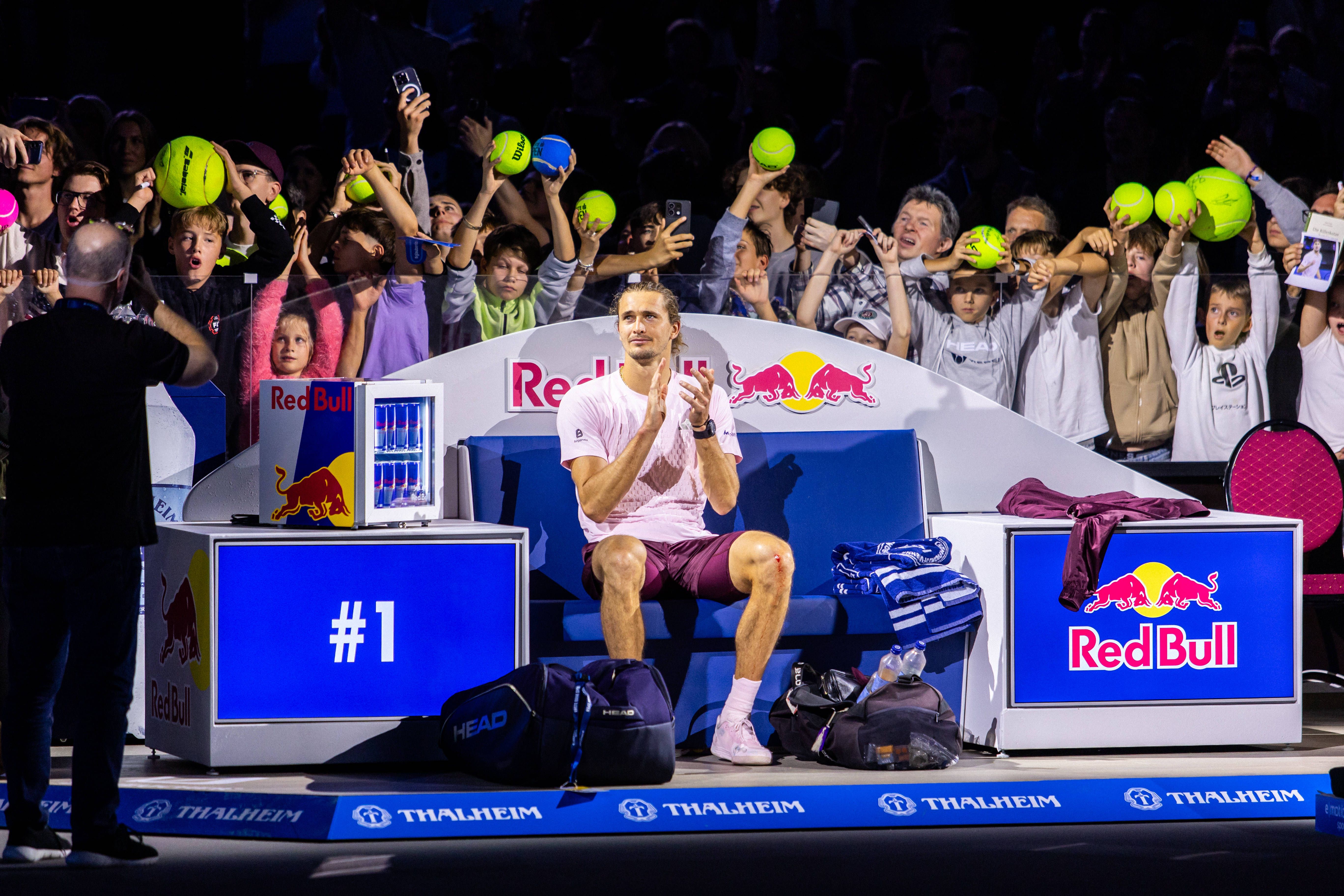 Alexander Zverev sitzt jubelnd auf der Spielerbank, Fans halten Bälle für Autogramme hoch.
