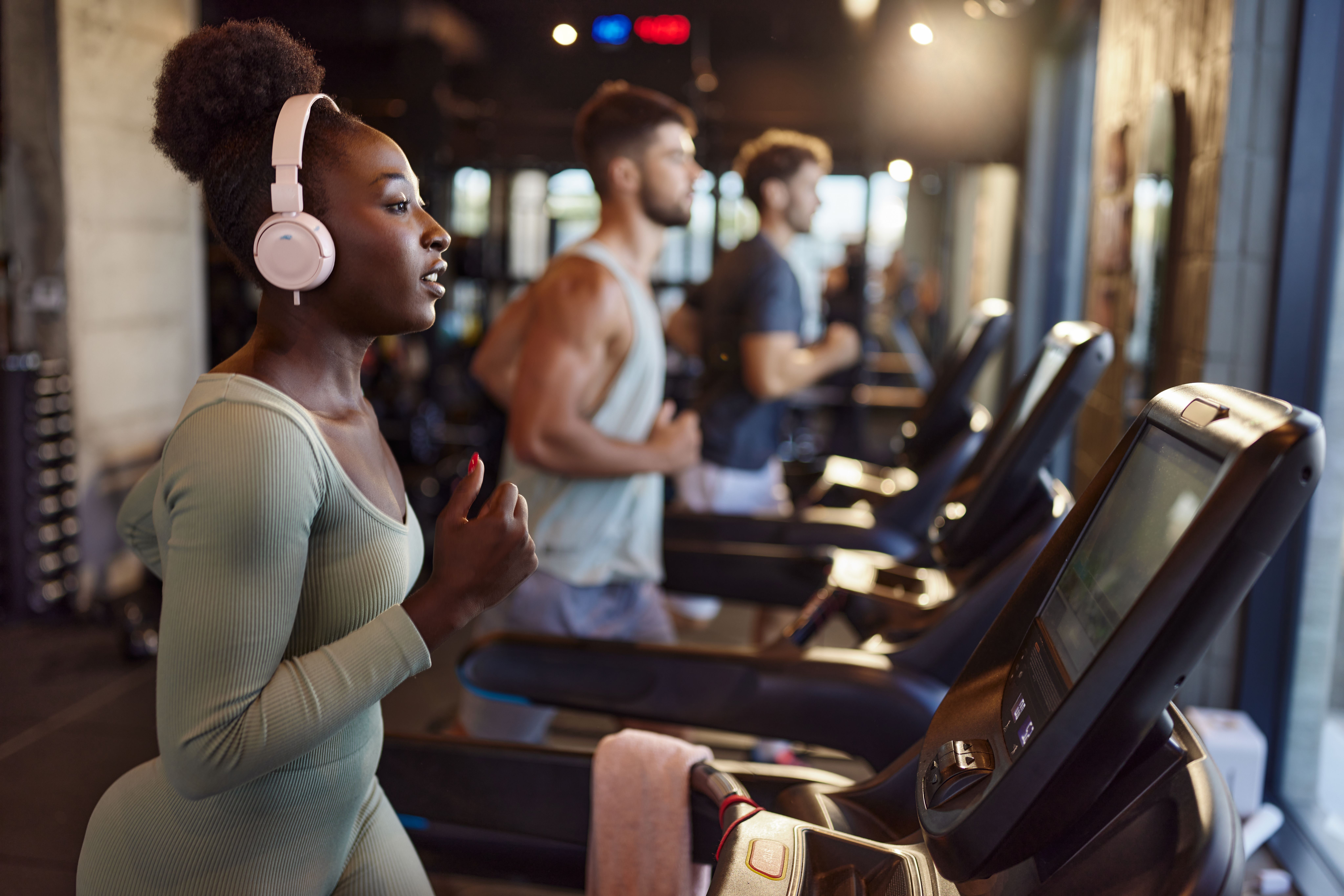Frau mit Kopfhörern läuft auf einem Laufband im Fitnessstudio, weitere Personen im Hintergrund.
