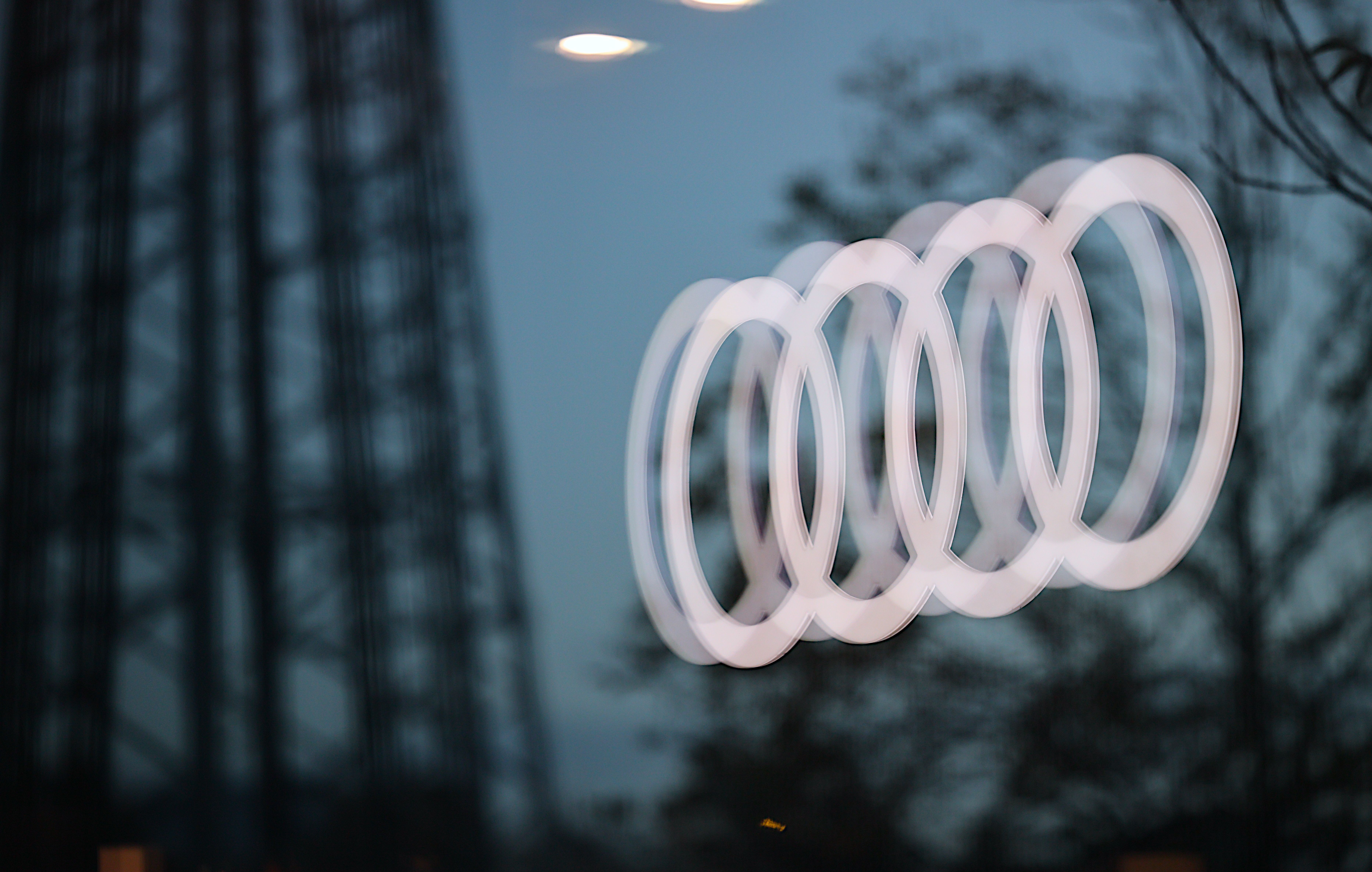 Verschwommenes Audi-Logo leuchtet vor einem Glasfenster mit Spiegelungen und dunkler Umgebung.
