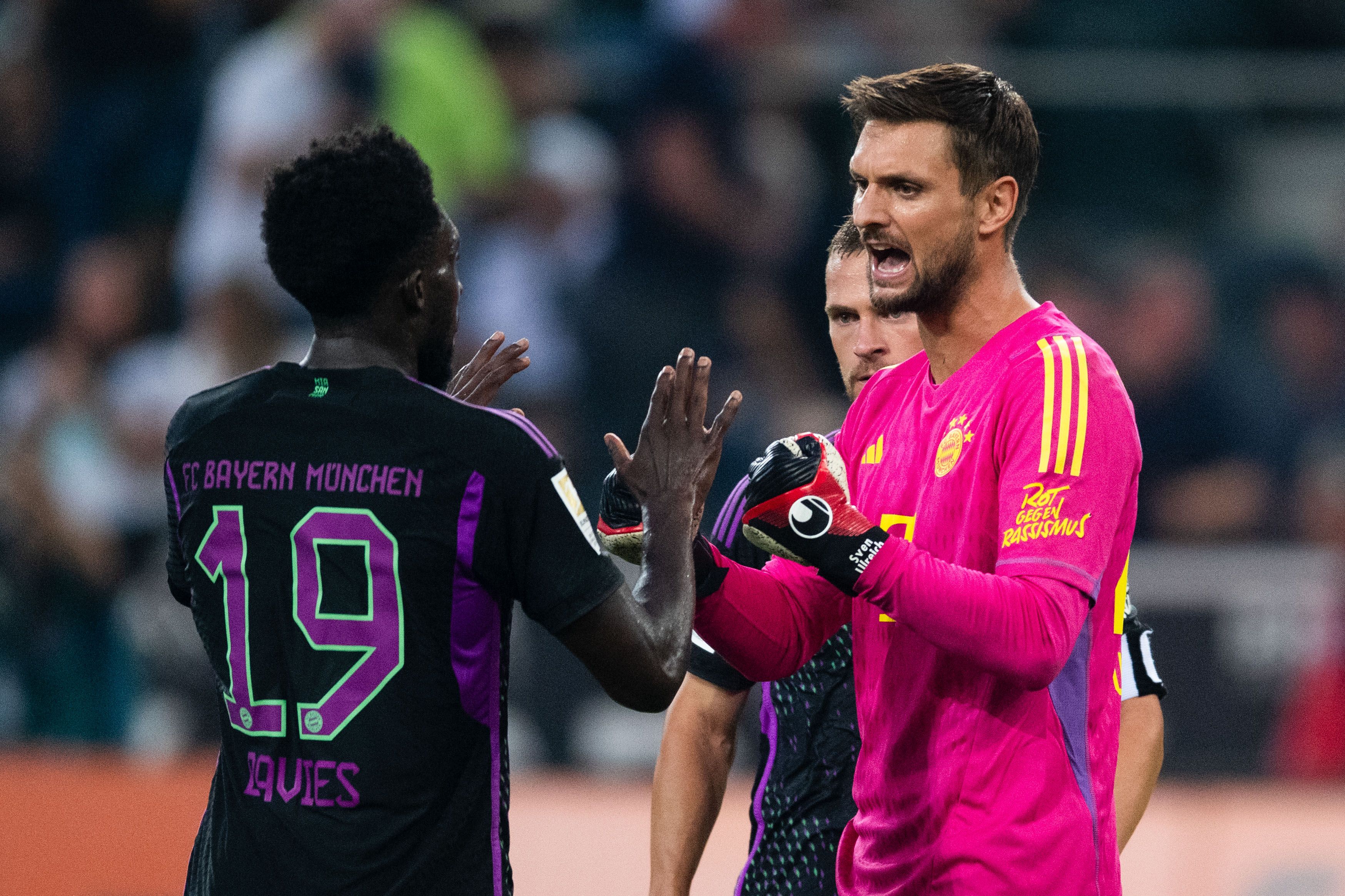 Bayern-Torhüter Sven Ulreich klatscht im pinken Trikot während des Spiels ab und gibt Anweisungen an Abwehrspieler Alphonso Davies.