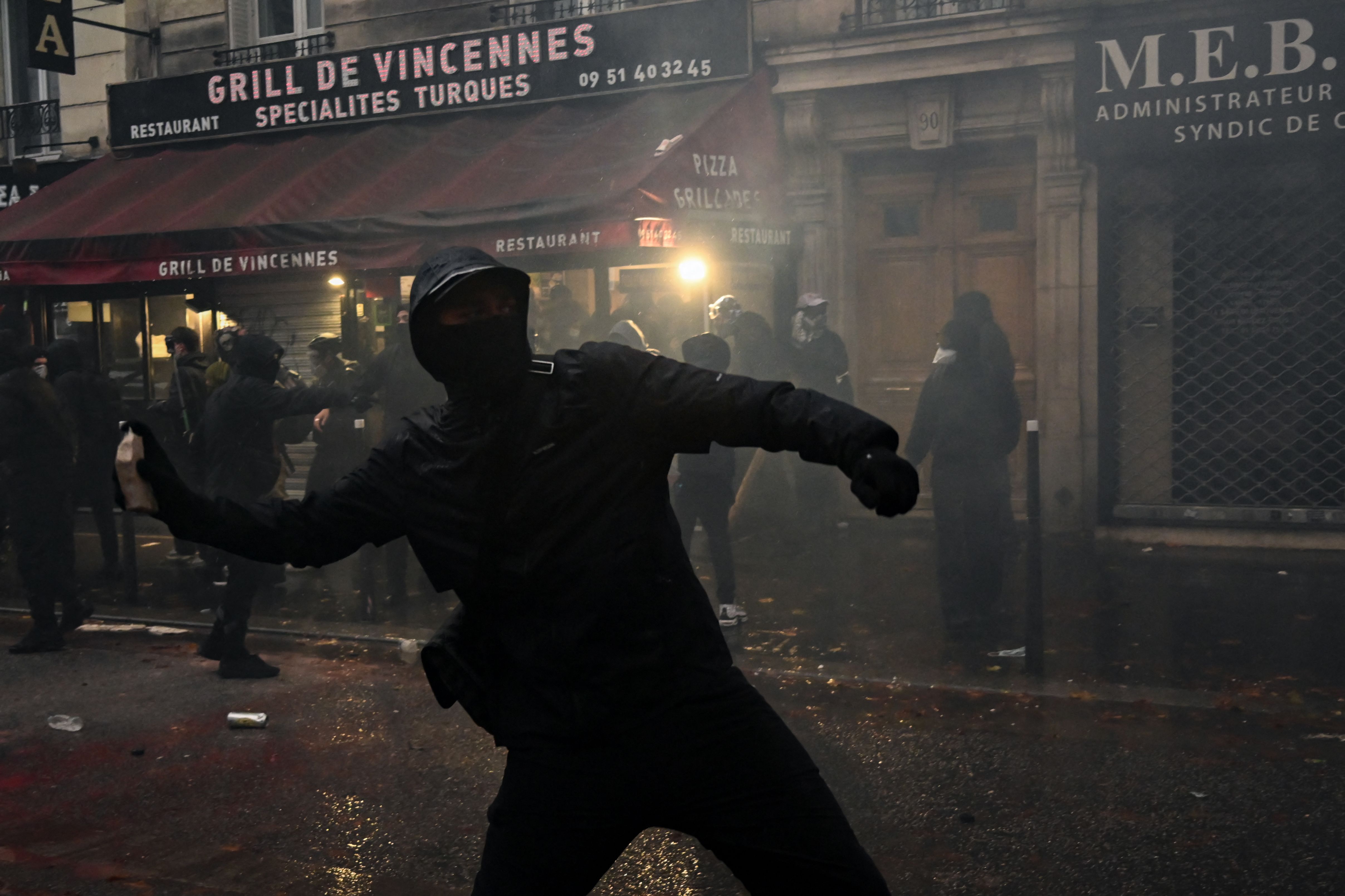 Vermummter Demonstrant wirft Gegenstand bei Protest in Paris vor Restaurant, im Hintergrund weitere Personen.