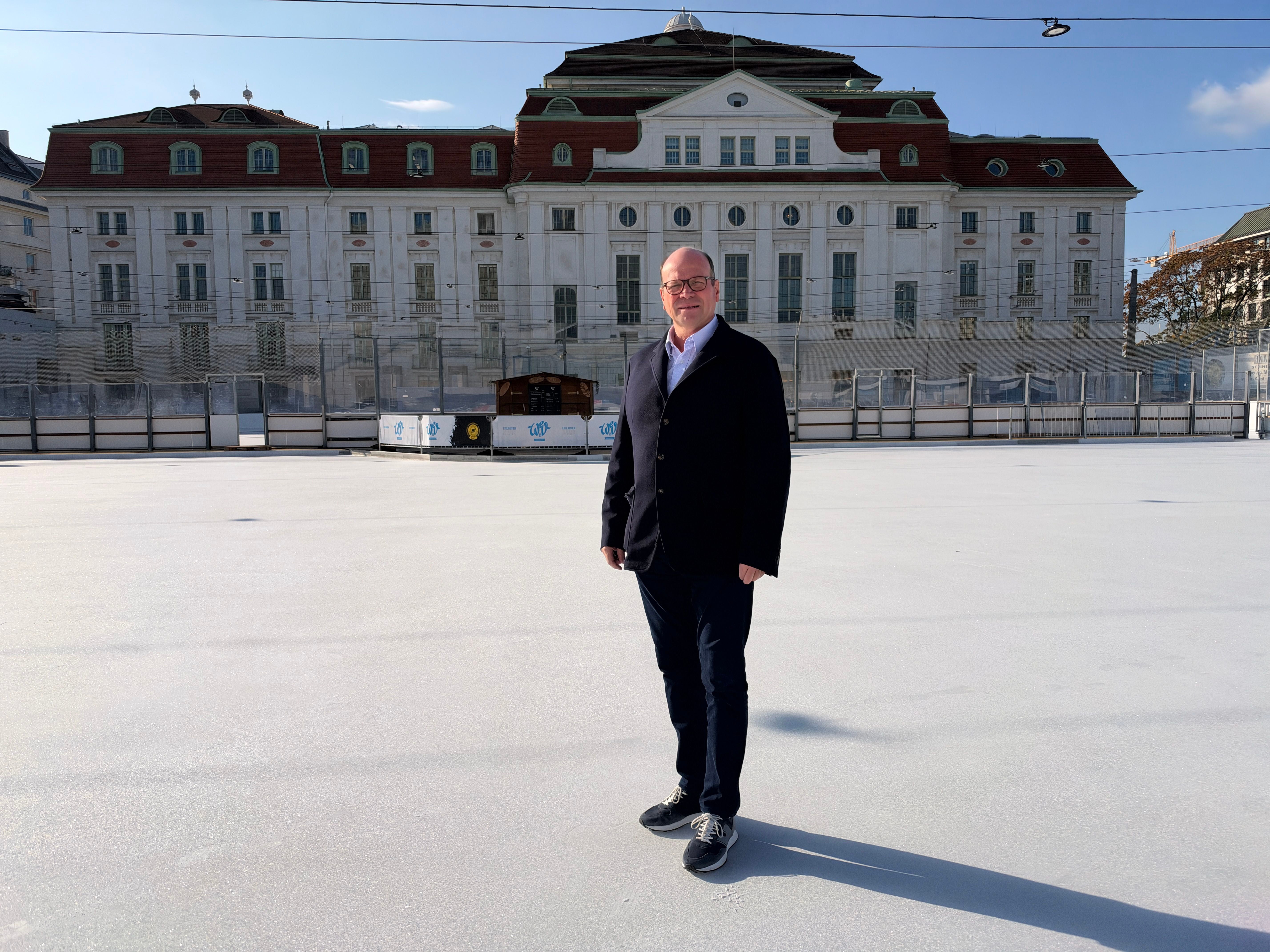 Ein Mann steht auf einer Eisfläche vor einem großen historischen Gebäude bei Sonnenschein.