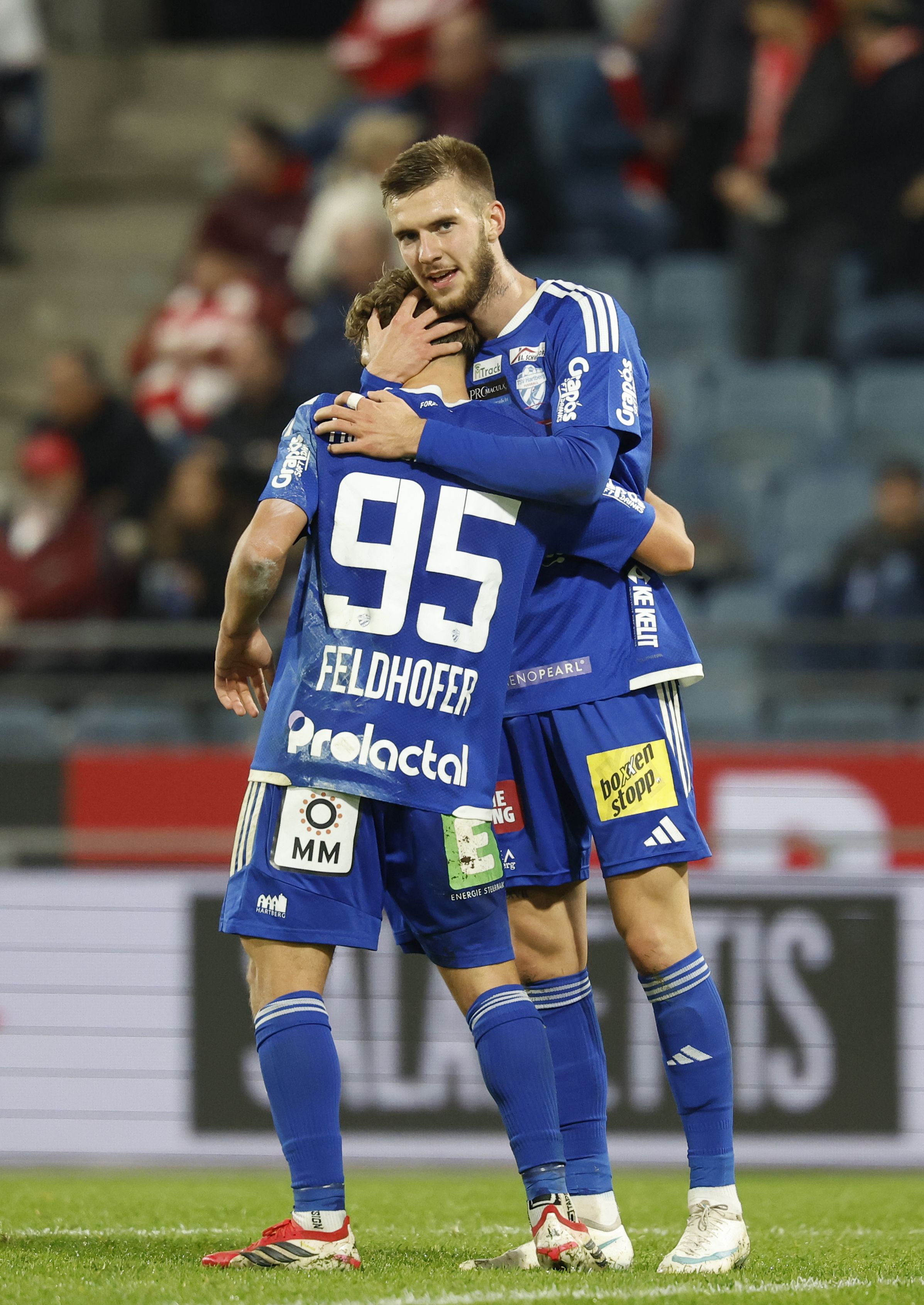 Hartberg-Torschütze Nikolaus Feldhofer im blauen Trikot mit der Rückennummer 95 umarmt jubelnd einen Teamkollegen Oliver Lukic auf dem Spielfeld.