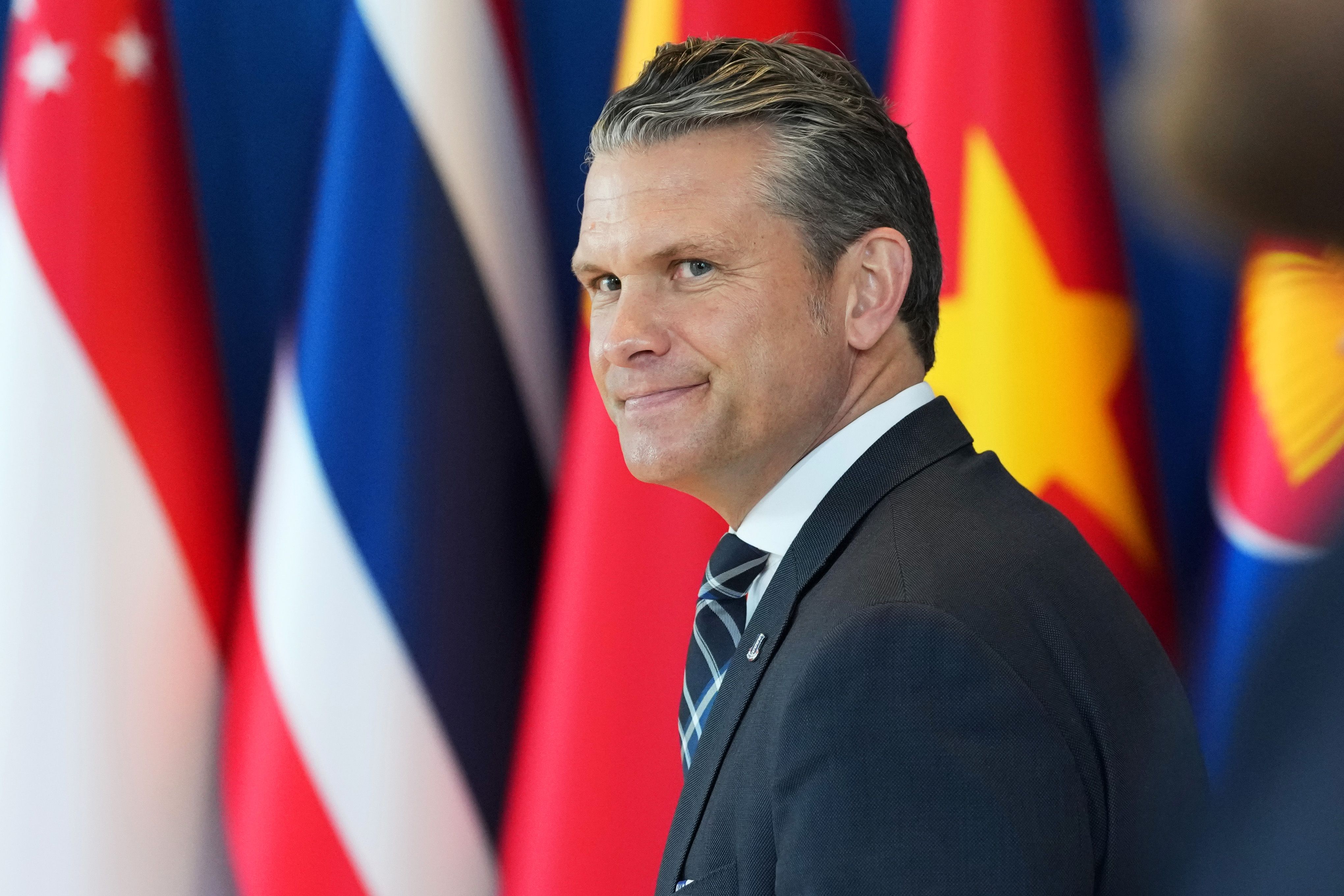 Ein Mann im Anzug lächelt vor unscharfen internationalen Flaggen im Hintergrund.