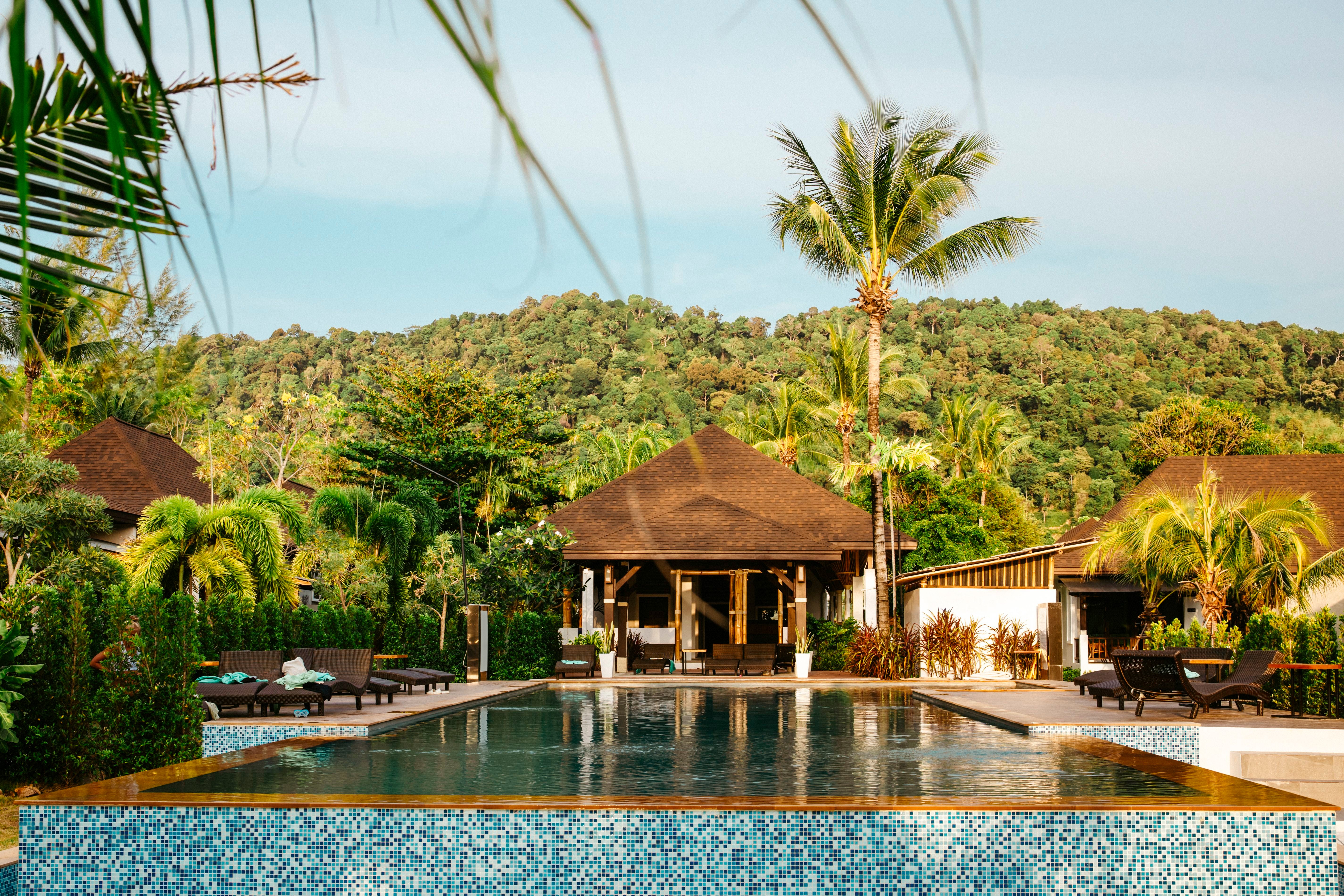 Pool mit Sonnenliegen und Palmen vor tropischem Hügel in einem Resort in Thailand.