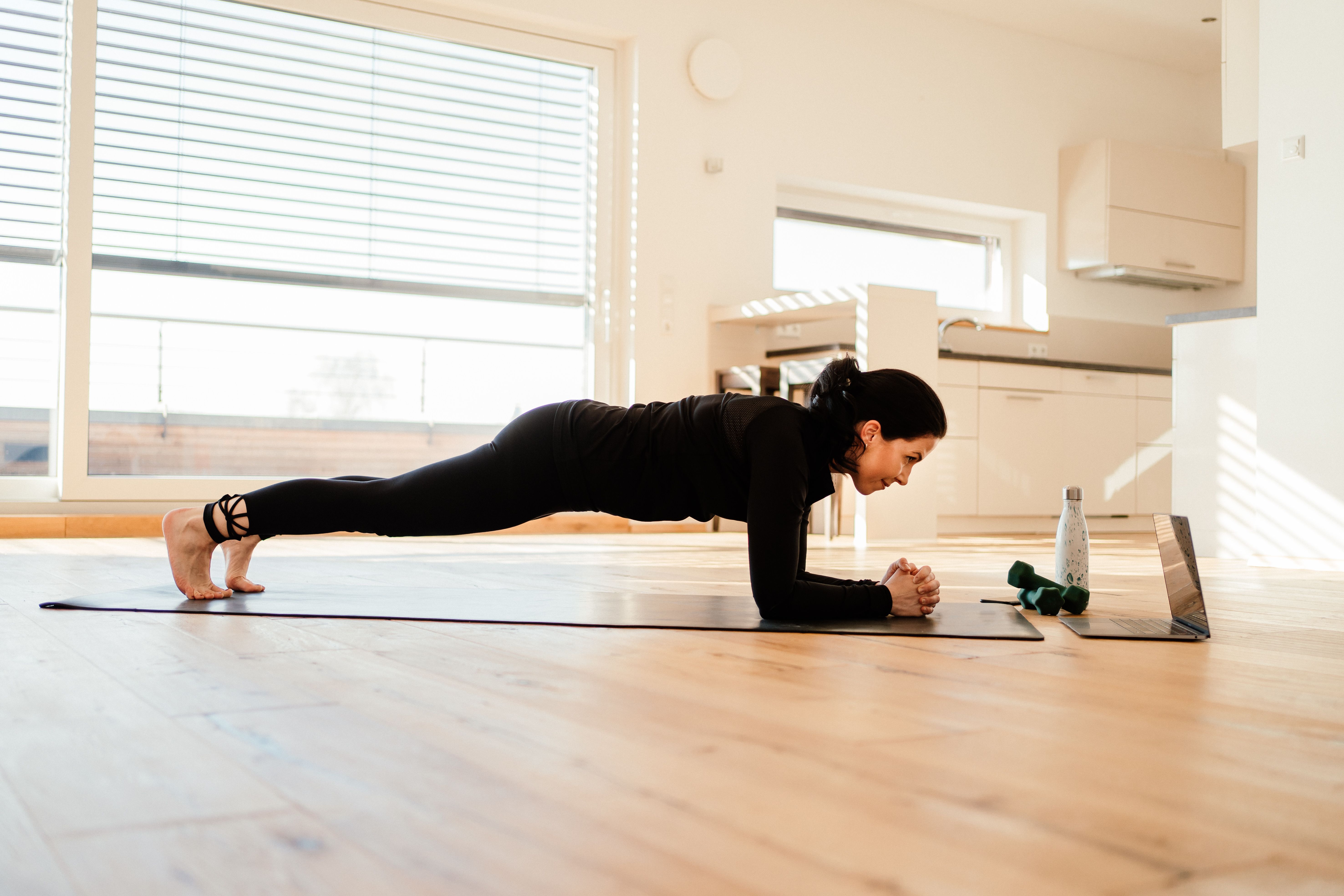 Frau hält Plank-Position auf Yogamatte in einem hellen Wohnzimmer mit Laptop und Hanteln.