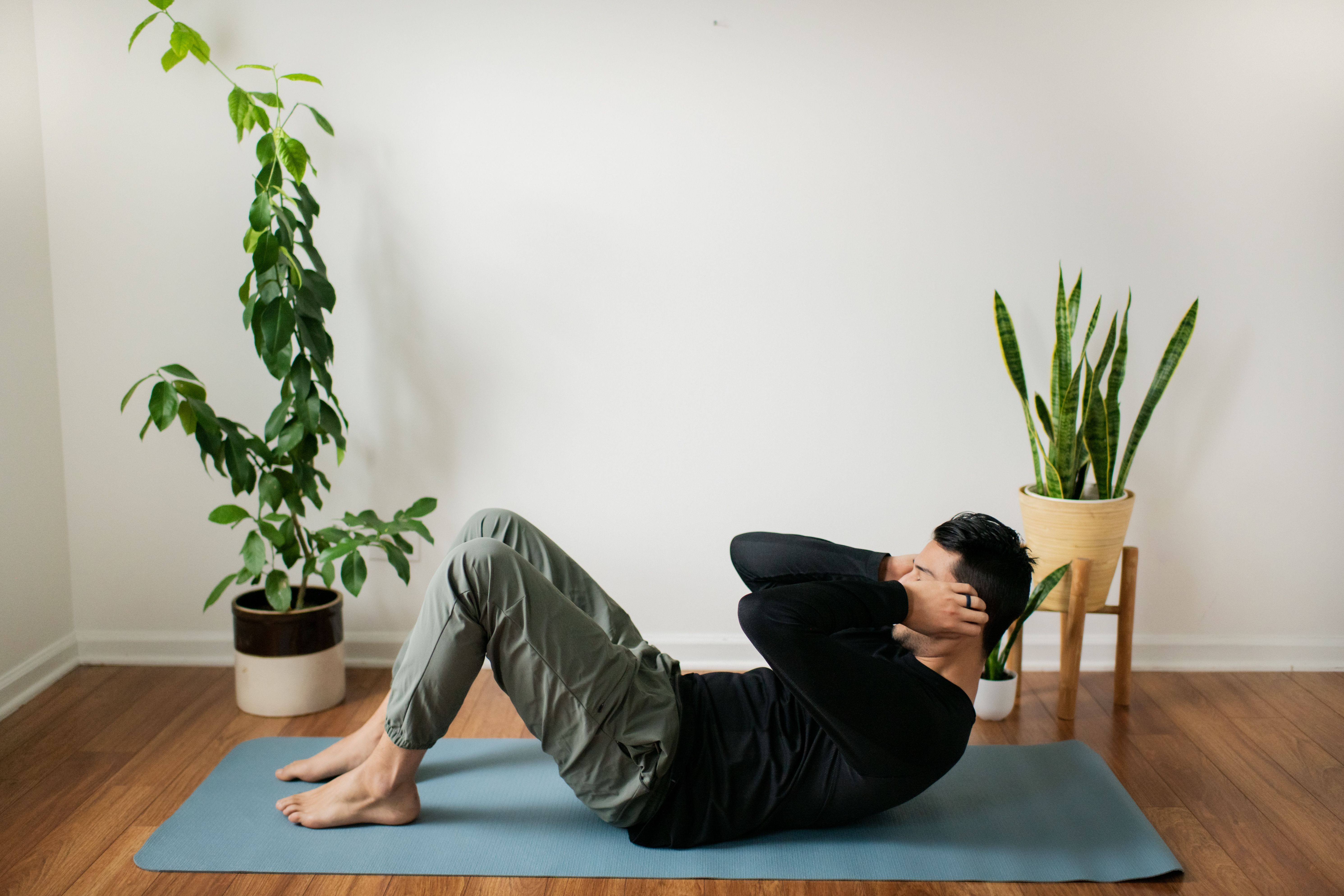 Mann macht Sit-ups auf einer Yogamatte im Wohnzimmer zwischen zwei Zimmerpflanzen.