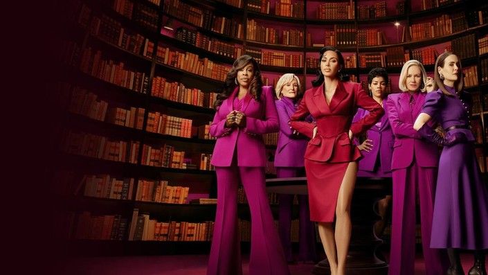 Sechs Frauen in pinker und lilafarbener Kleidung stehen in einer Bibliothek mit vielen Büchern.